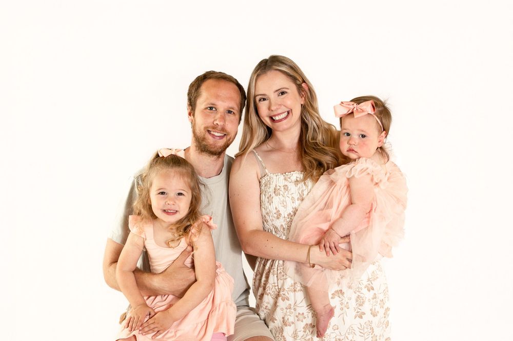 Happy family of four posing together, parents holding two young daughters in matching pink outfits against white background.