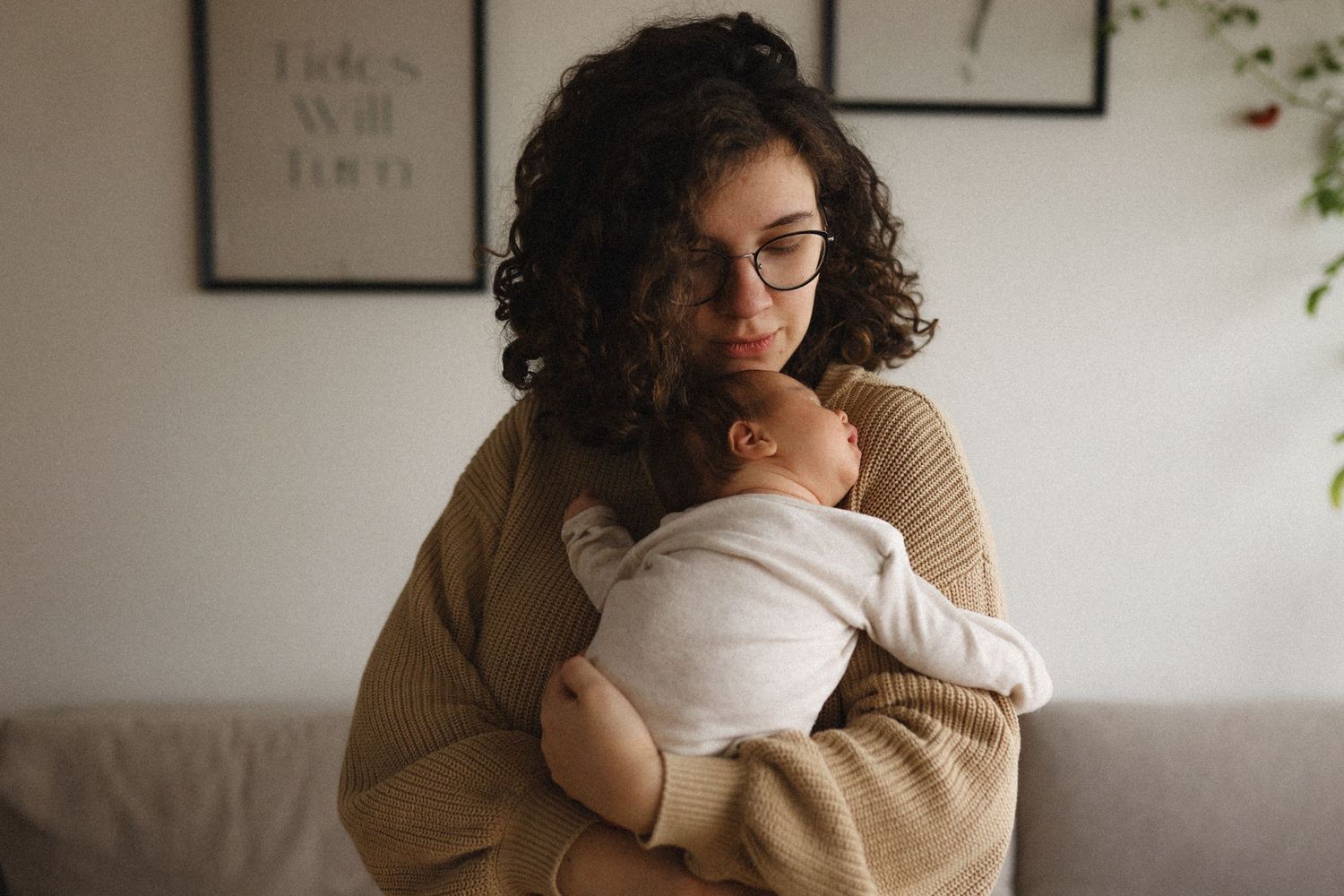 A person with curly hair wearing a beige sweater cuddles a swaddled baby while sitting on a couch.