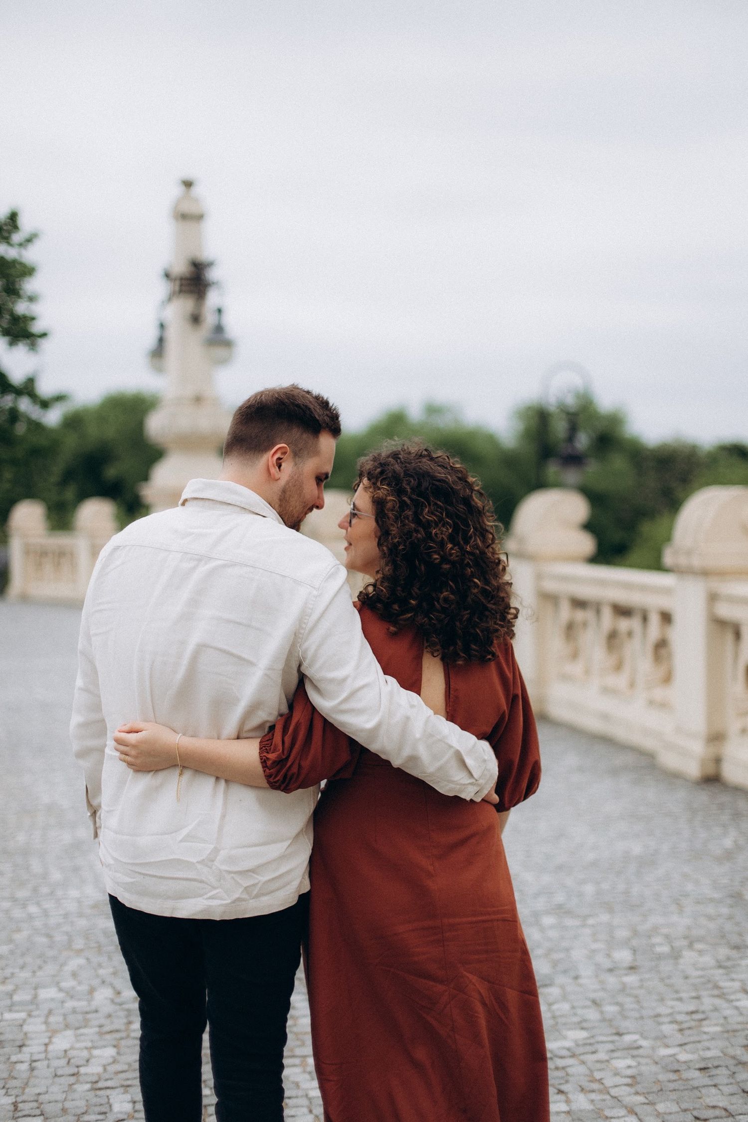 Couple in coordinating white and burgundy outfits embrace while walking on cobblestone path near ornate architecture.