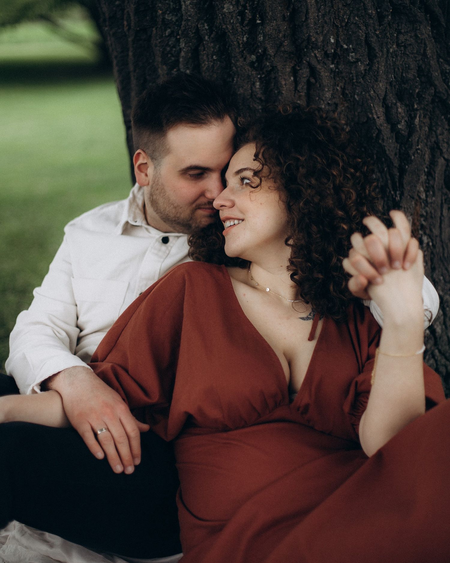 A romantic couple embraces against a tree trunk in a garden, with one wearing a red dress and the other in a white shirt.