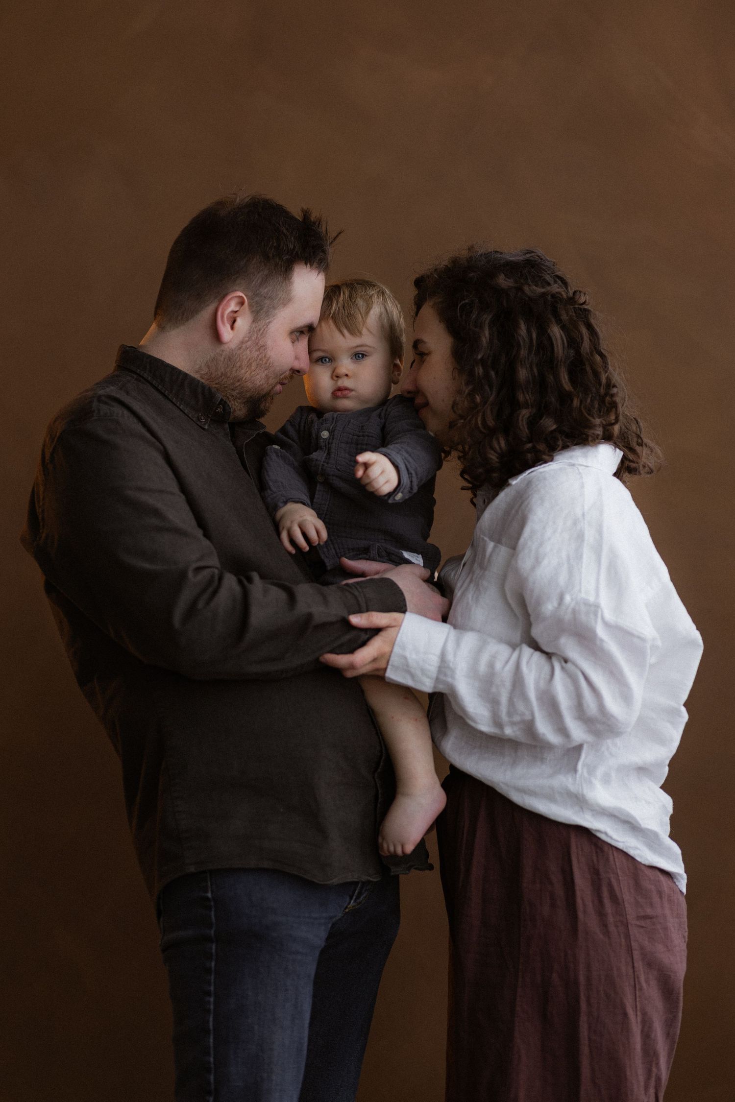 A small family sharing an intimate moment together against a brown studio backdrop.