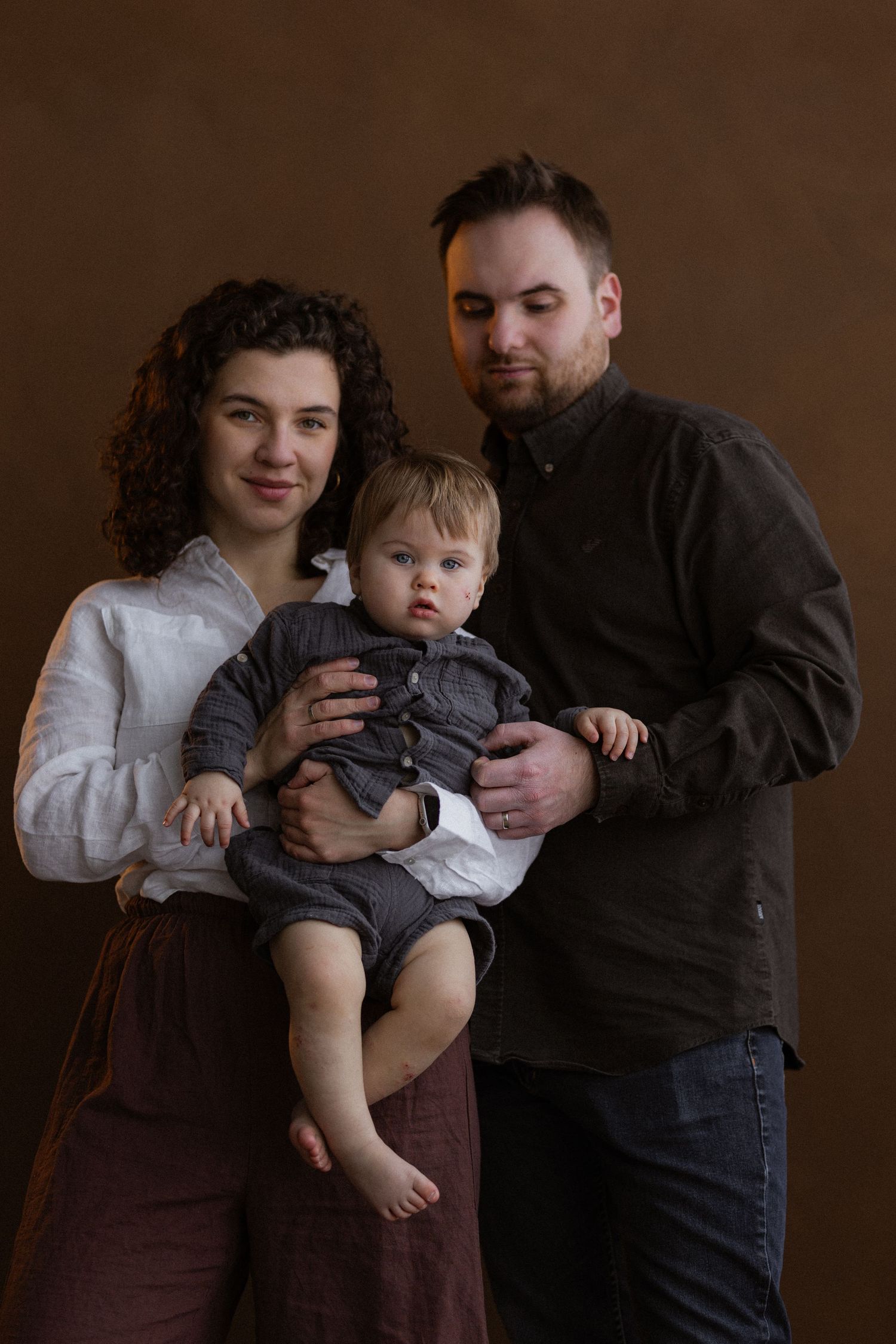 Professional family portrait in moody lighting against brown backdrop with parents holding young toddler in grey outfit.