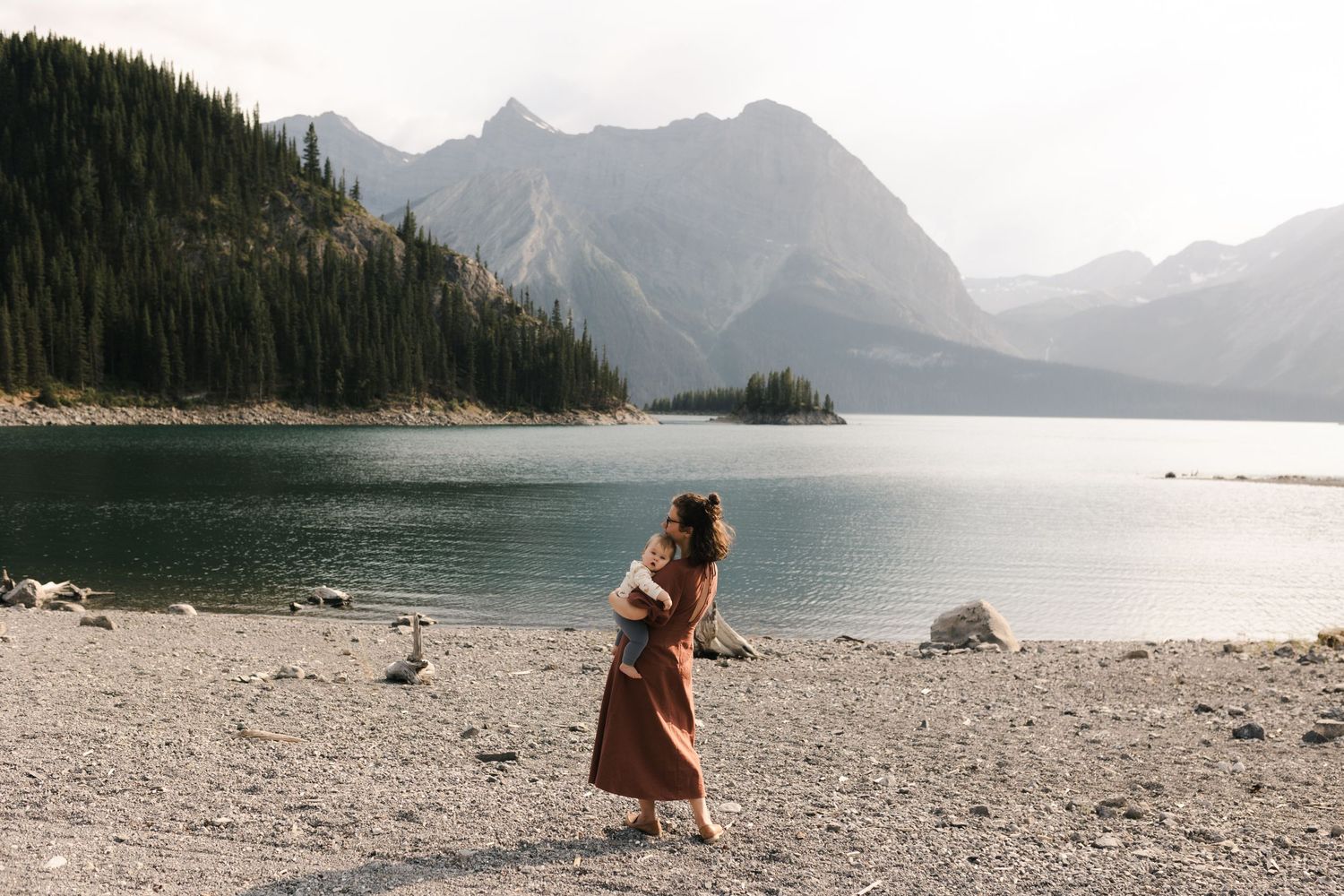 Someone in a long brown coat walks along a rocky beach with mountains and a serene lake in the background.
