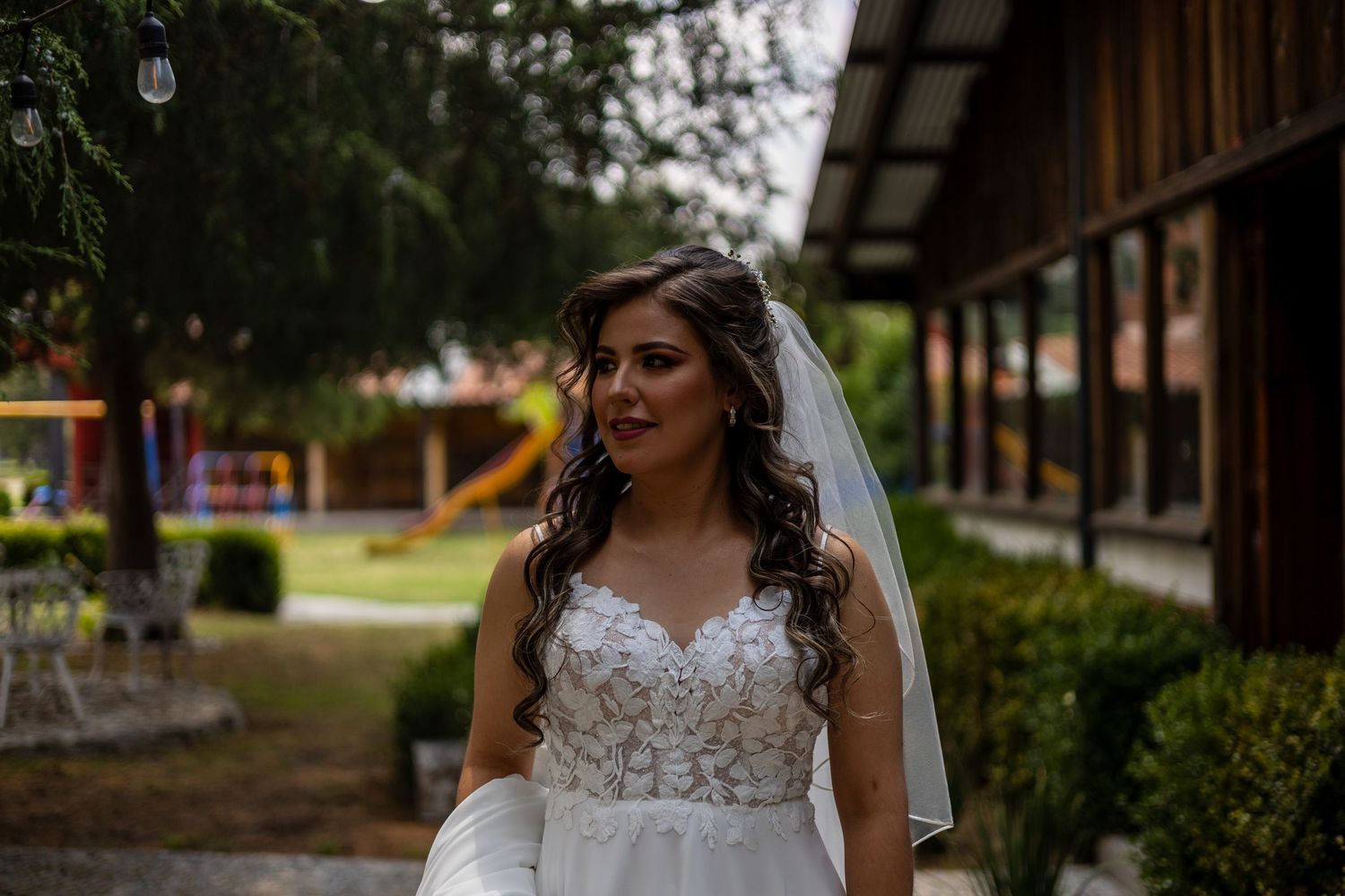 Bride in lace wedding dress poses in front of rustic wooden building.
