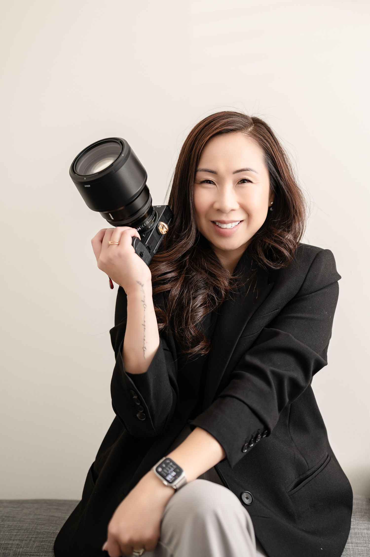 A photographer wearing an off-shoulder beige sweater smiles while holding a professional camera in white and grey setting.