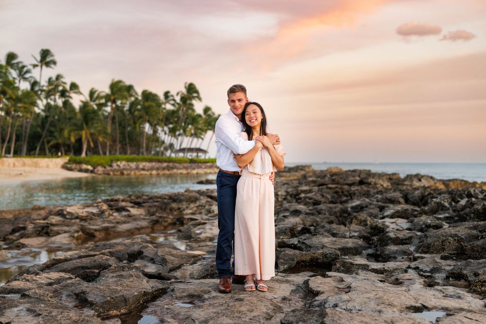 Couple embracing on rocky Hawaiian beach at sunset with palm trees and pink sky backdrop.