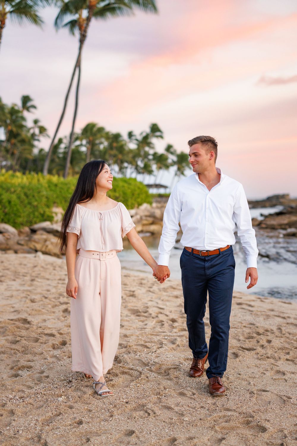 Couple holding hands walking on a sandy beach at sunset with palm trees in the background.