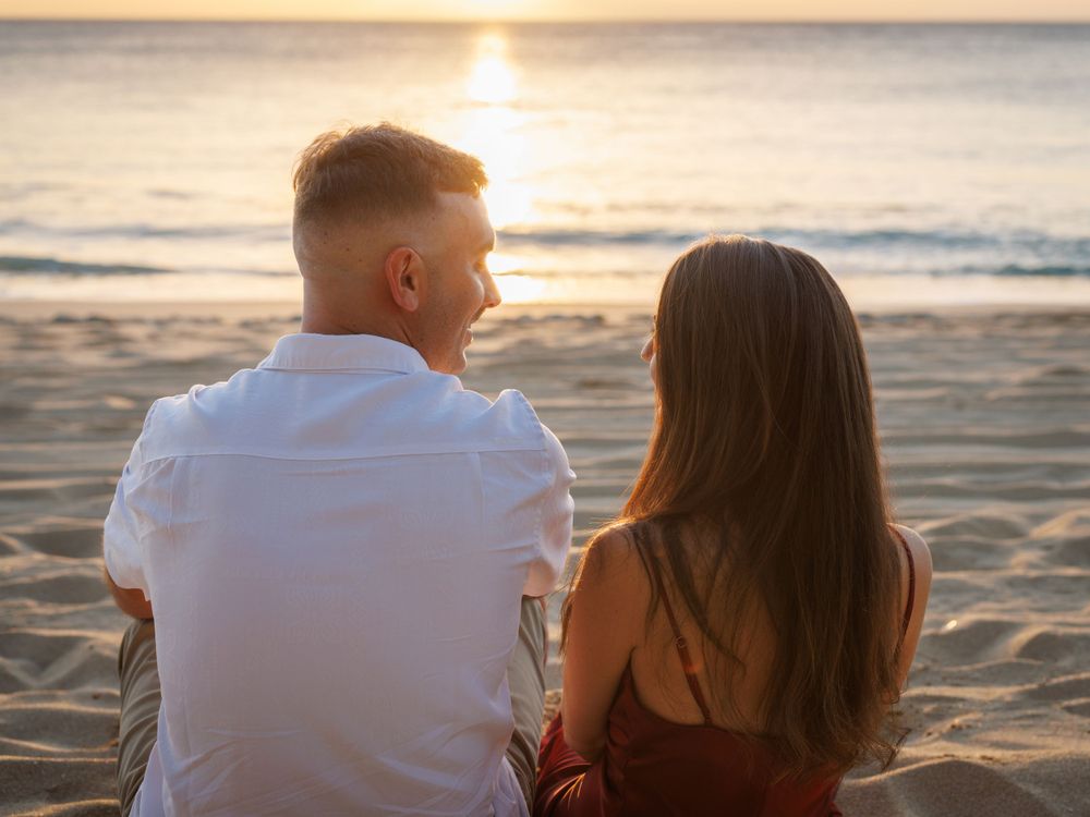 Couple sitting together on a sandy beach watching a romantic golden sunset over the ocean.