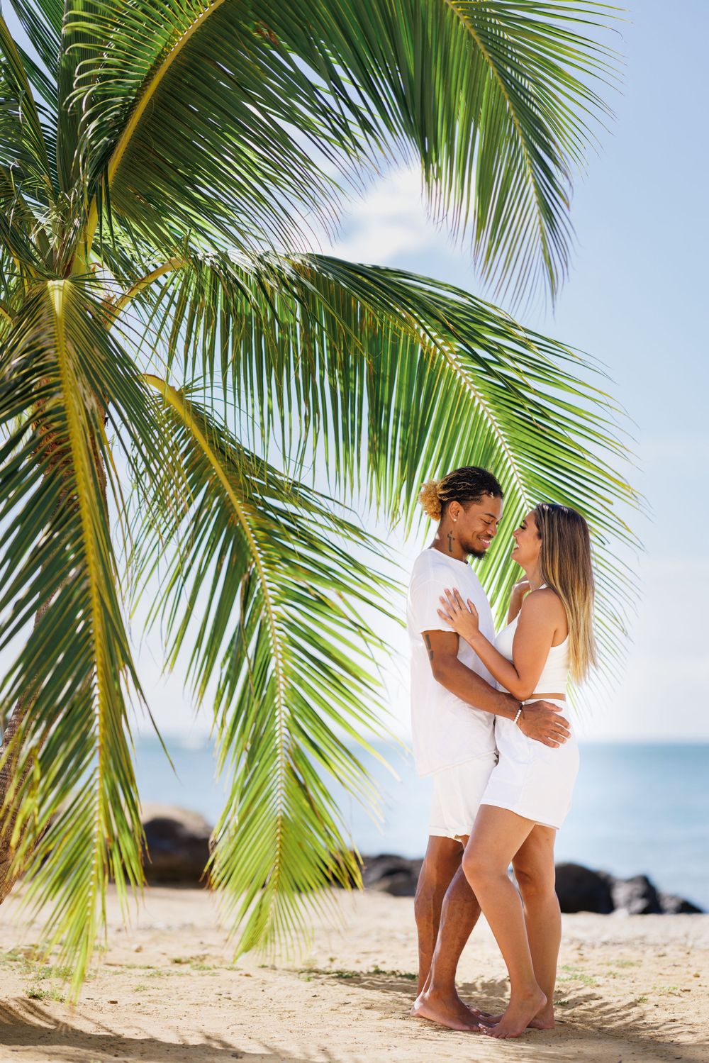Romantic couple embracing under a tropical palm tree on a sandy beach with blue ocean in the background.