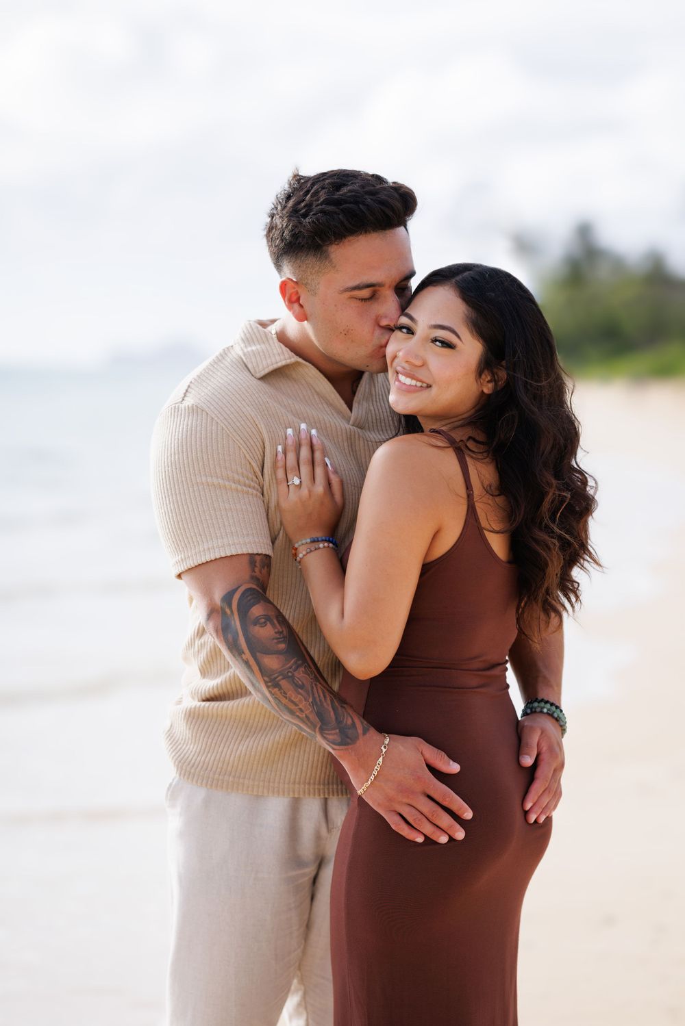 Couple embracing on a beach, man kissing woman's cheek, she smiles showing engagement ring, tropical setting.