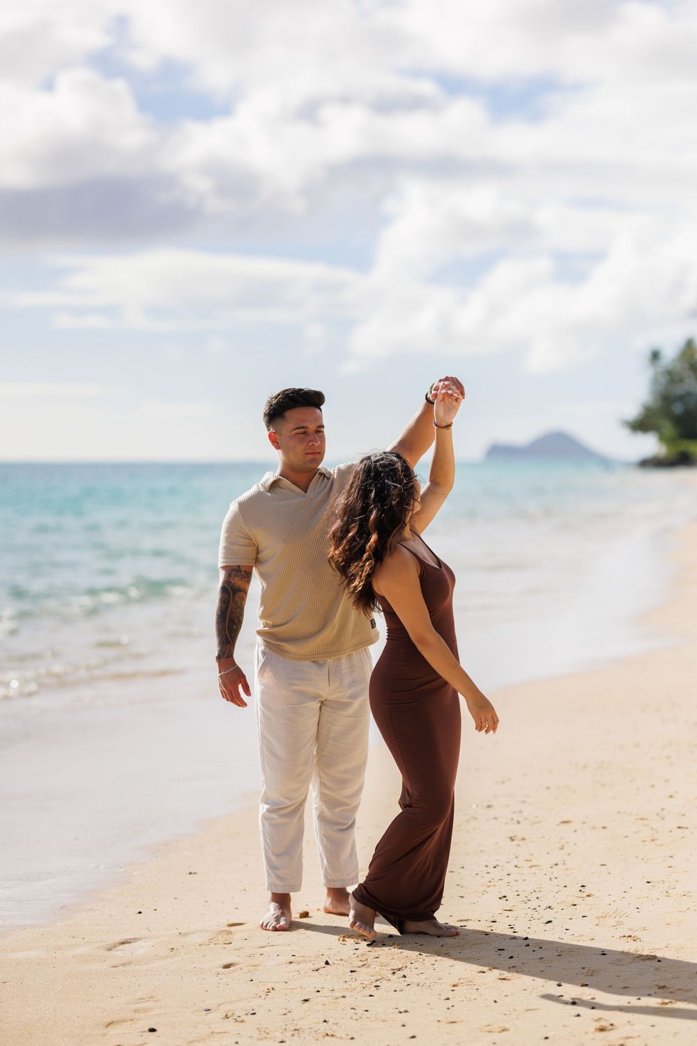 Couple dancing on a tropical beach, man in beige shirt and white pants, woman in brown dress, turquoise ocean backdrop.