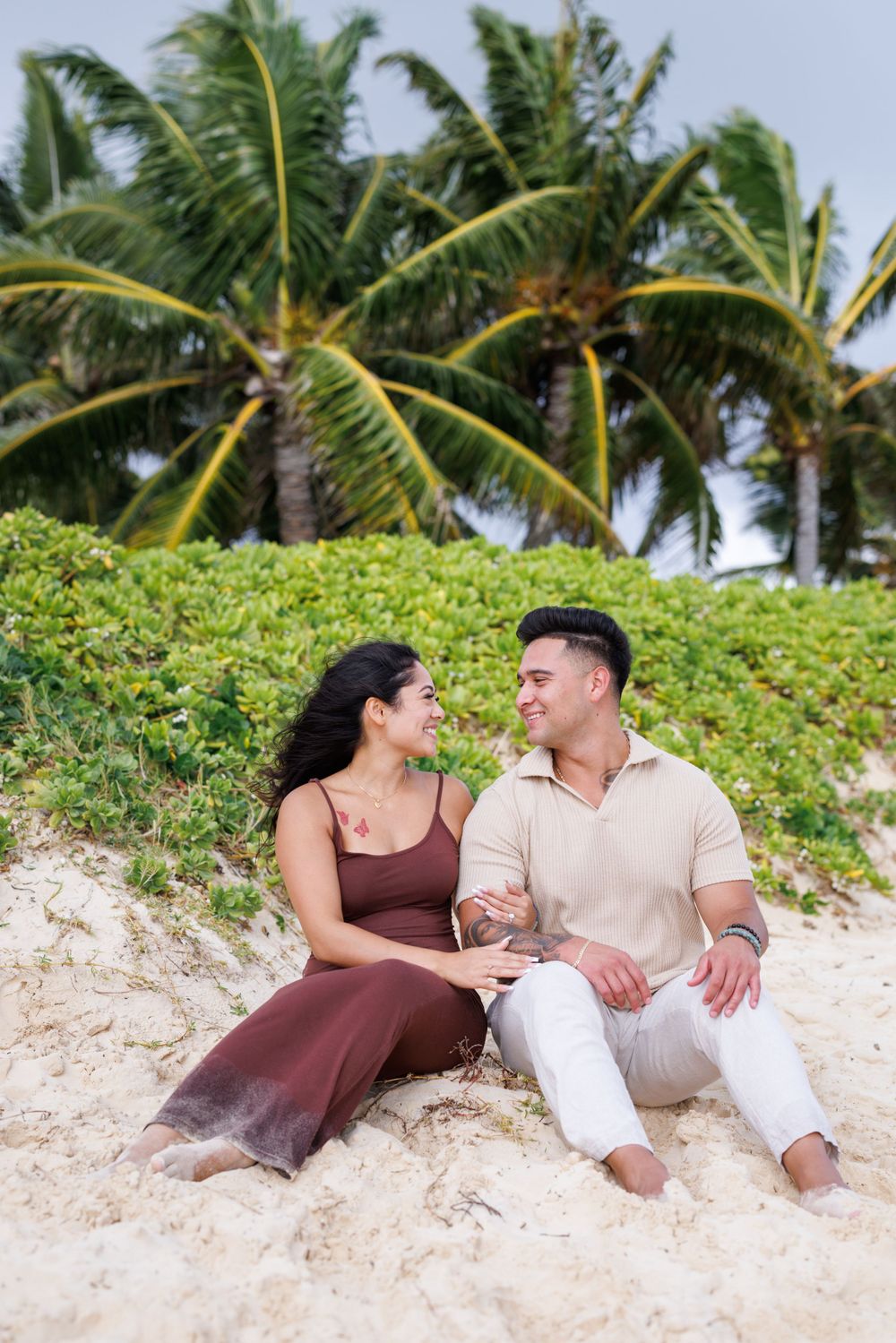 Couple sitting together on sandy beach smiling at each other with tropical palm trees and greenery behind them.