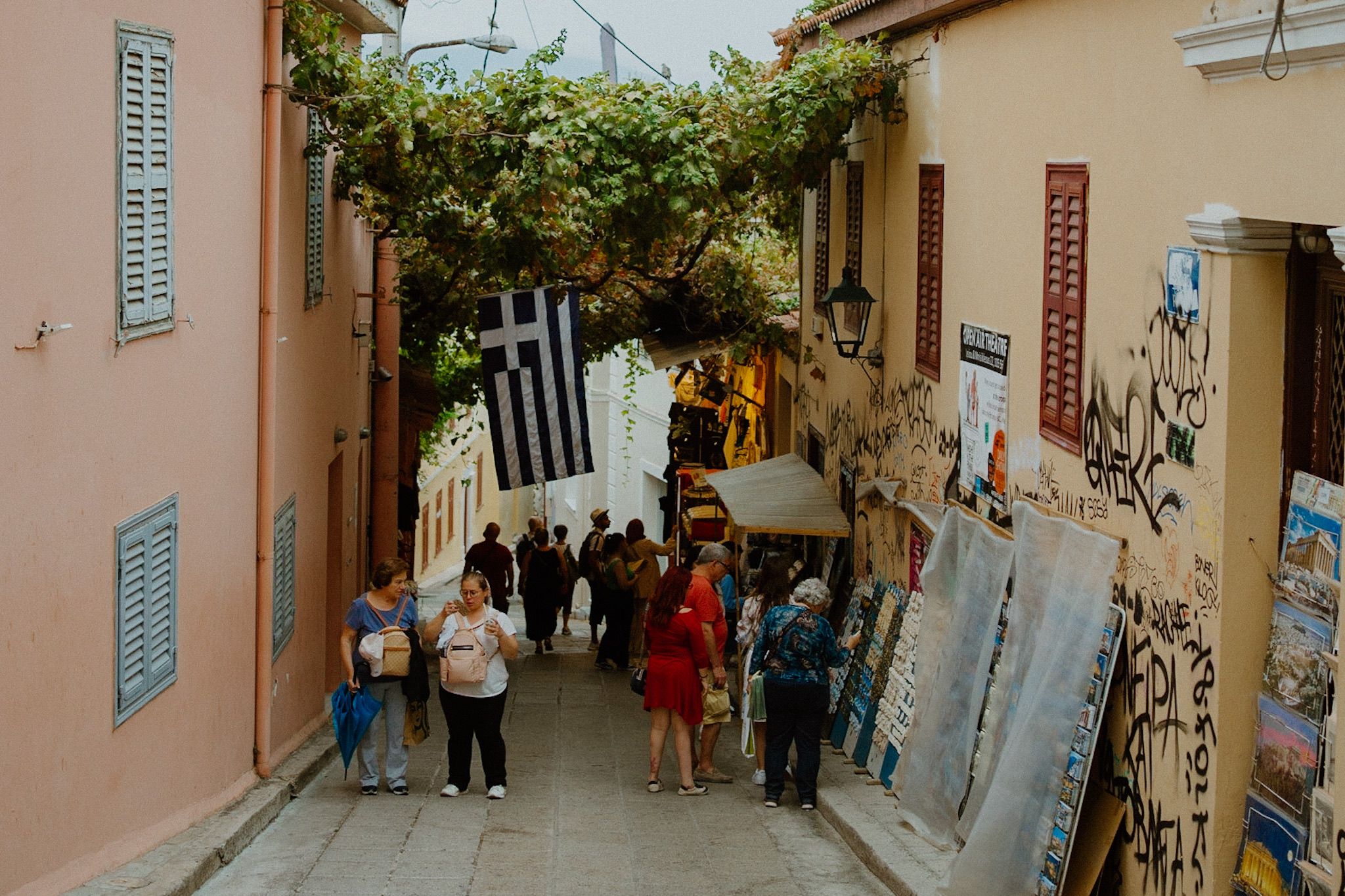 Tourists walk down a narrow cobblestone street between colorful buildings in Athens, Greece with blue shutters and graffiti.