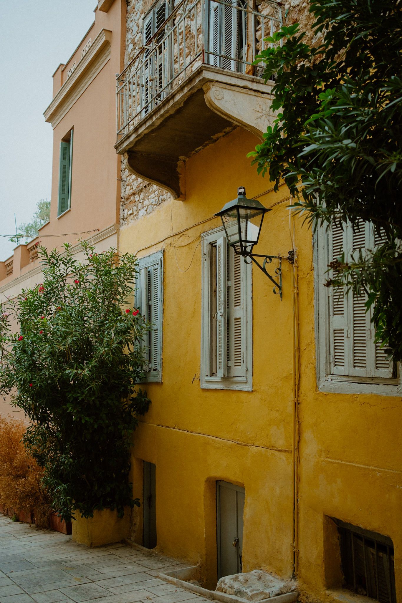 A traditional yellow Mediterranean building with blue shutters and an ornate iron balcony on a quiet street.