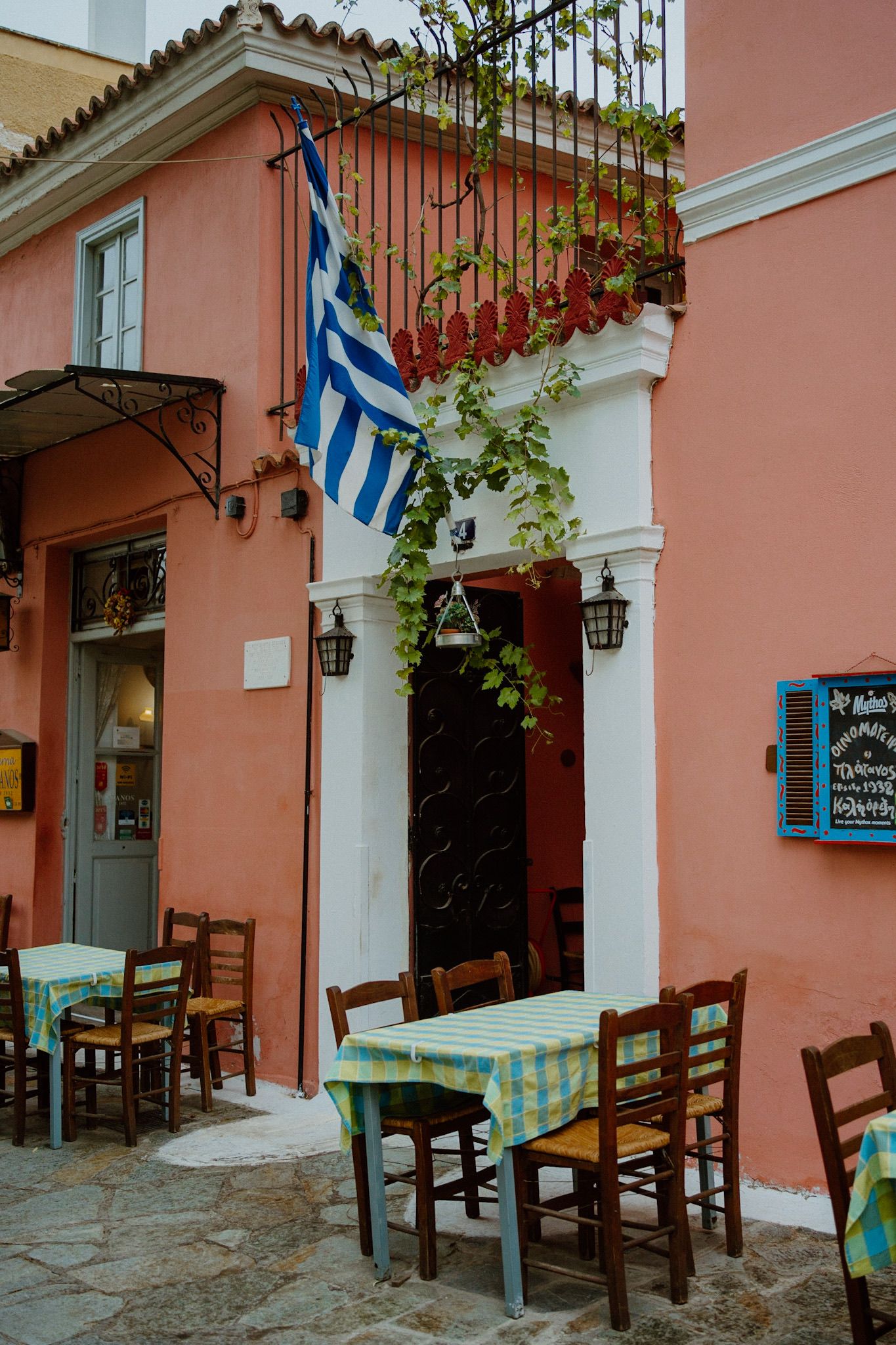 Traditional Greek taverna exterior with pink walls, hanging flag, and outdoor seating with checkered tablecloths.
