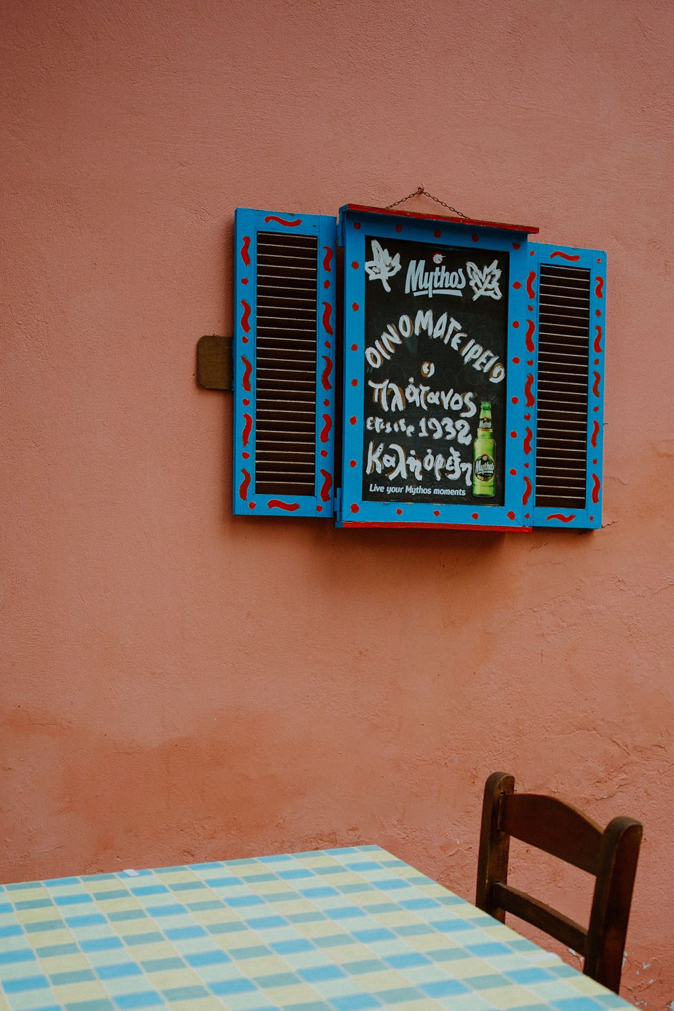 Blue window shutters with menu text on a terracotta wall above a checkered tablecloth and wooden chair.