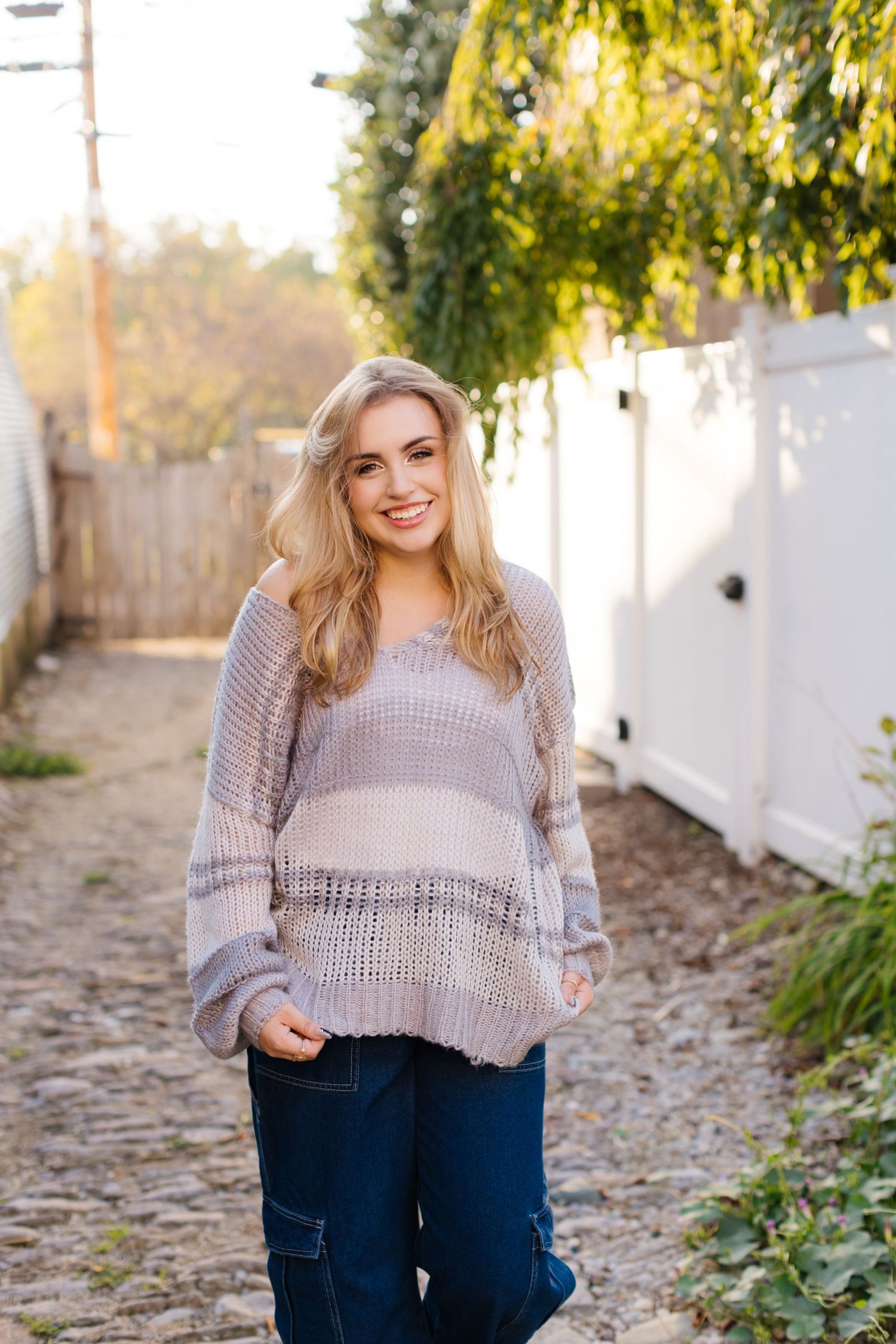 A person wearing a cozy striped knit sweater and jeans stands on a rustic path beside a white fence on a sunny day.