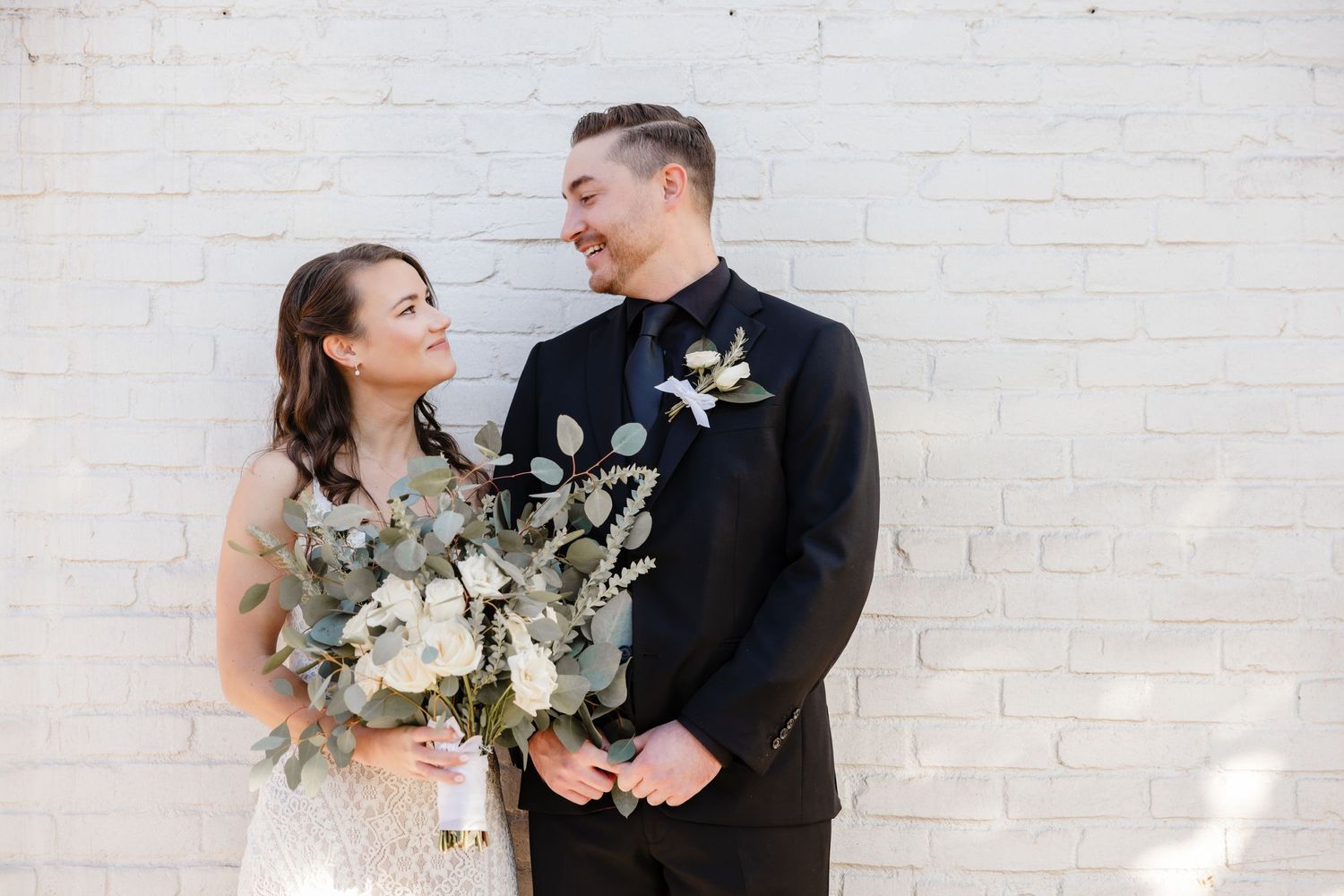 Wedding couple poses against white brick wall with elegant white and green botanical bouquet.