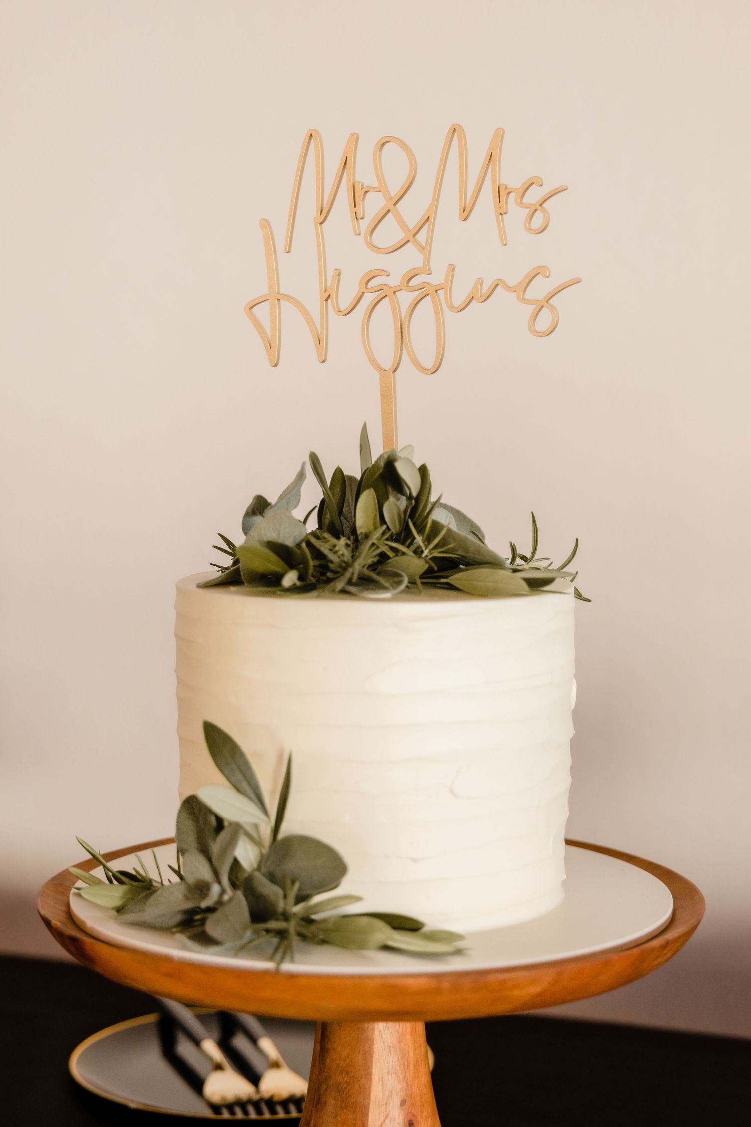 Simple white wedding cake decorated with eucalyptus leaves and gold 'Mr & Mrs' topper on wooden cake stand.