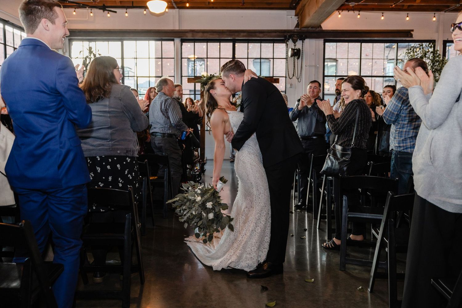 Wedding guests dance and celebrate together in a rustic industrial venue with large windows and exposed brick walls.