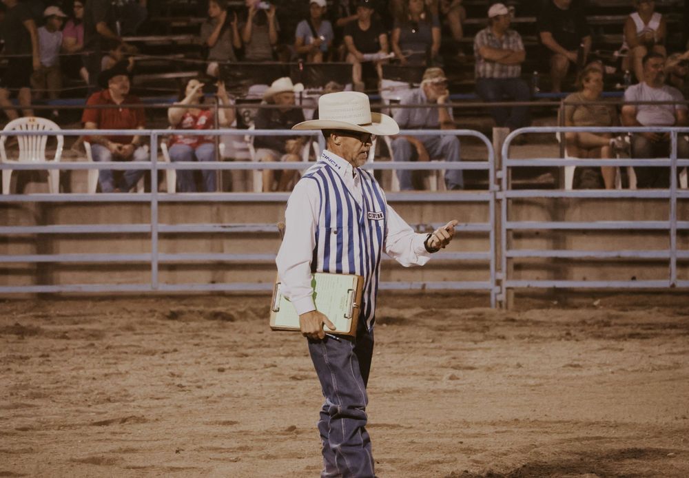 Rodeo official in striped shirt stands in sandy arena with spectators in background.