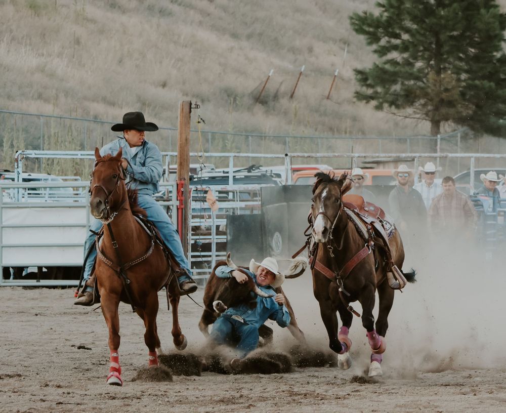 Cowboys competing in team steer wrestling event at dusty outdoor rodeo arena.