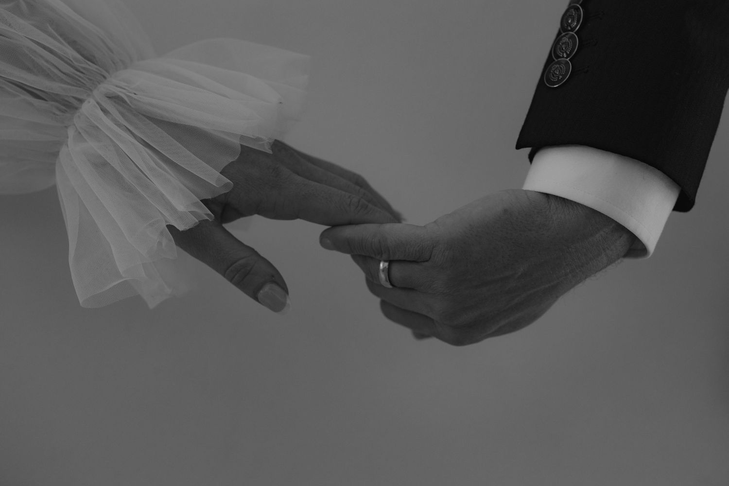 Black and white close-up photo of two hands reaching towards each other, one wearing a suit sleeve and one in a tulle veil.