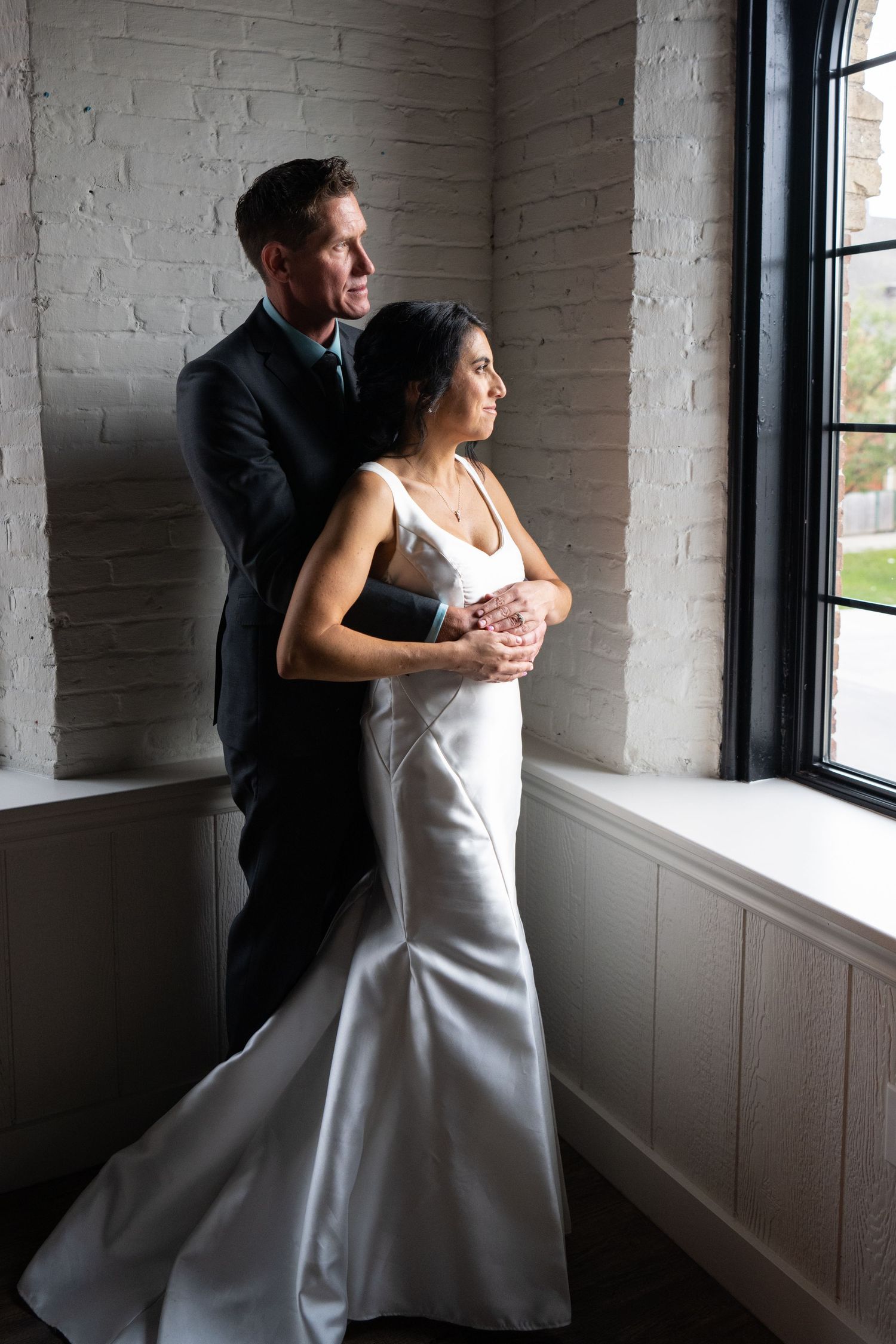 An intimate wedding moment by a bright window with a couple dressed in elegant formal attire. The Pie Eyed Monk