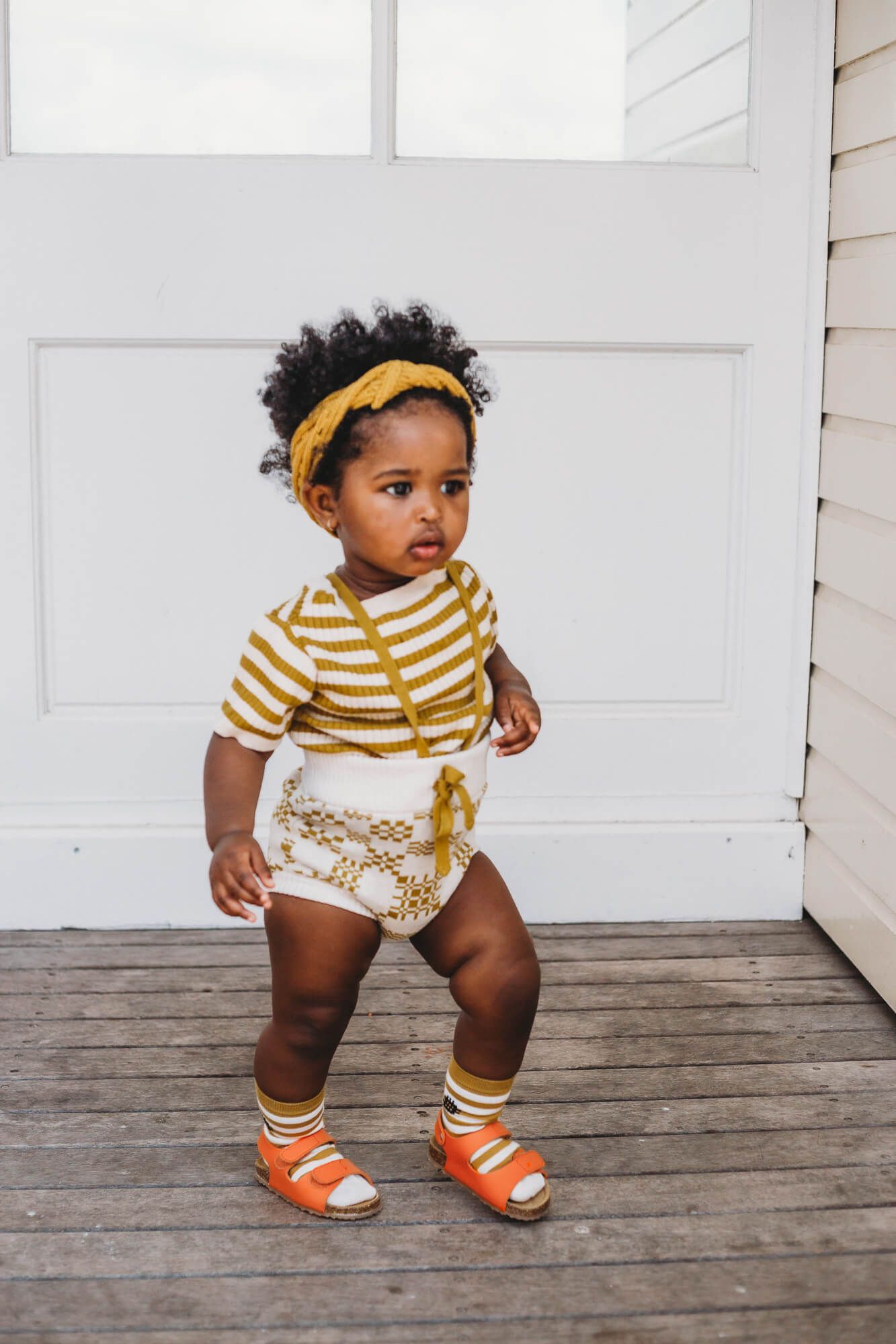 Small child in yellow striped top, yellow headband, patterned shorts, and orange socks stands on wooden deck.