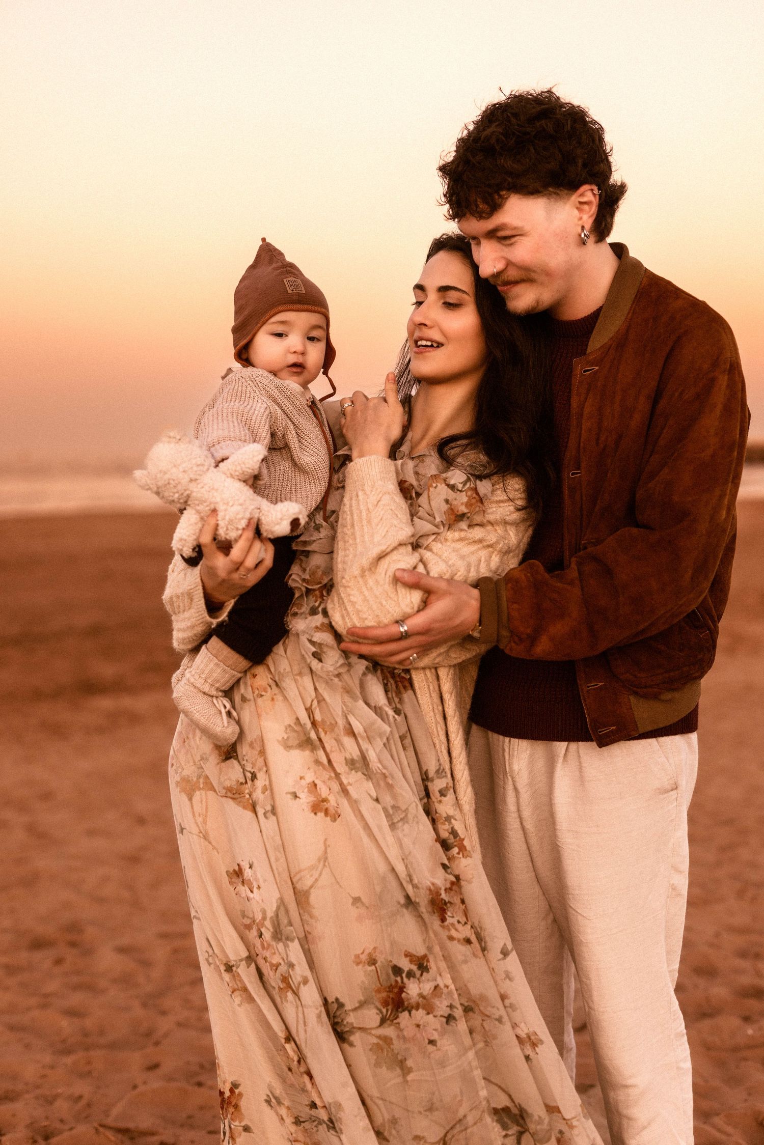 A family walks together on a hillside at sunset,with the mother wearing a floral dress and father holding their baby with photographer Javea