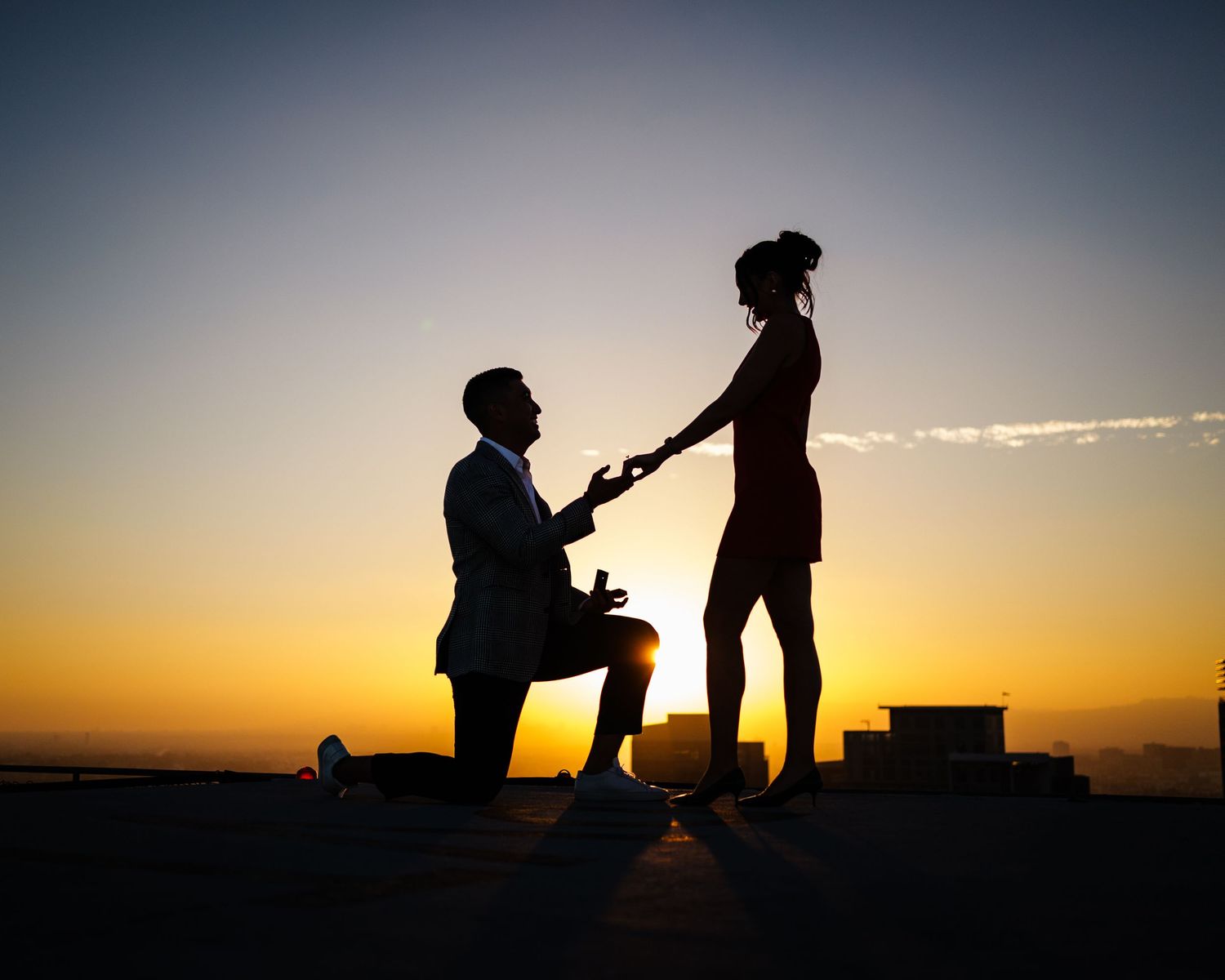 A silhouetted marriage proposal scene at sunset with an orange sky and glowing horizon in the background.