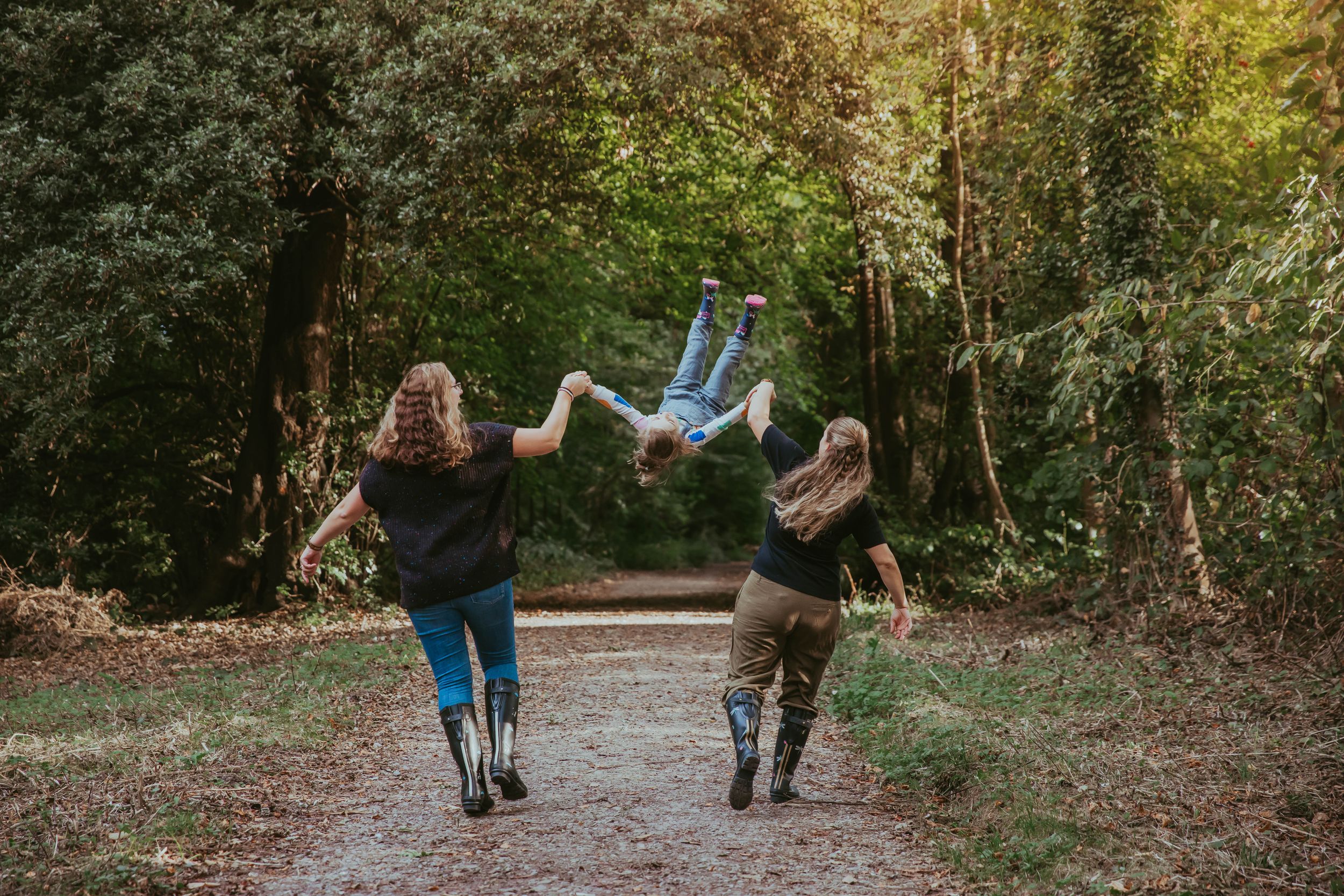 Two people walking down a sunlit forest path, playfully lifting a child upside down between them.