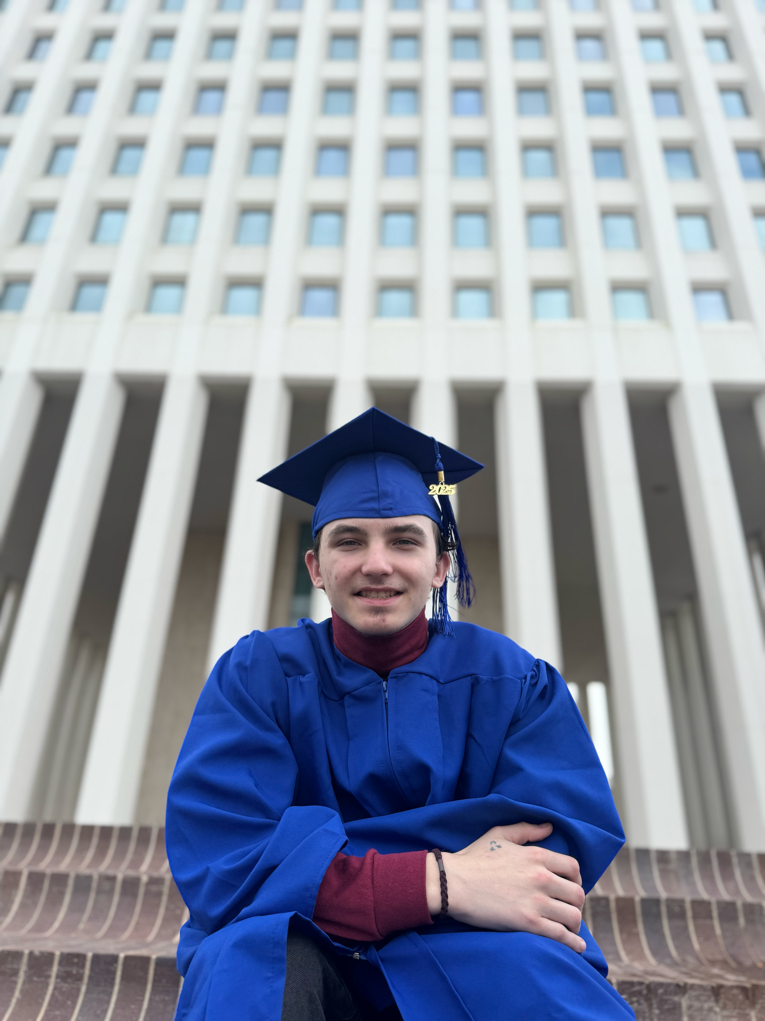 Graduate in blue cap and gown sits on steps in front of modern building with distinctive columns and windows.