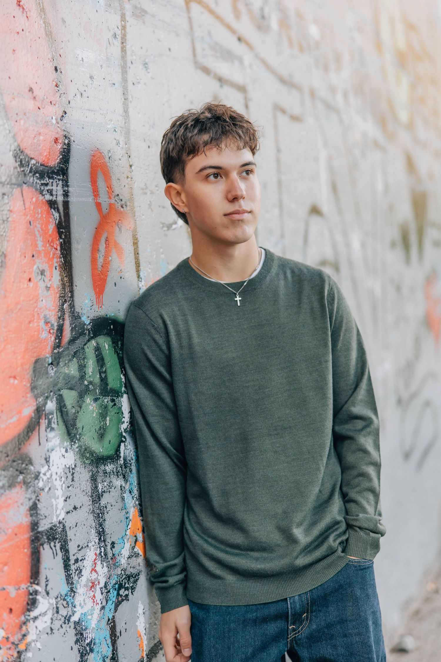 Teen boy in green sweatshirt and jeans leaning against a colorful graffiti wall outdoors.
