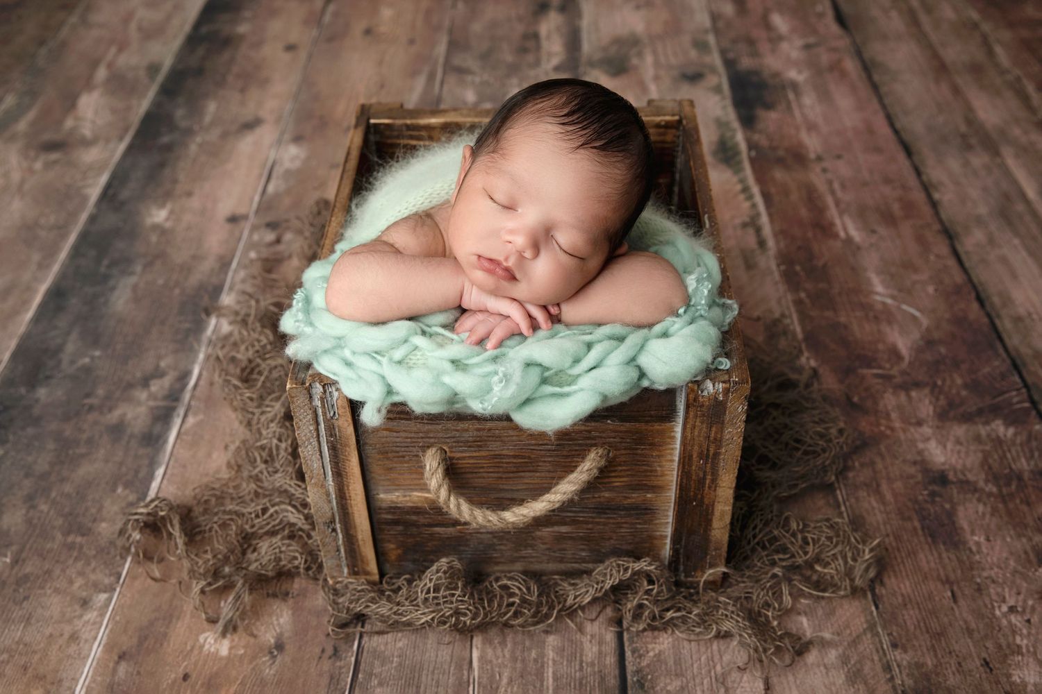 Sleeping newborn in rustic wooden crate with teal fabric on wooden floor.