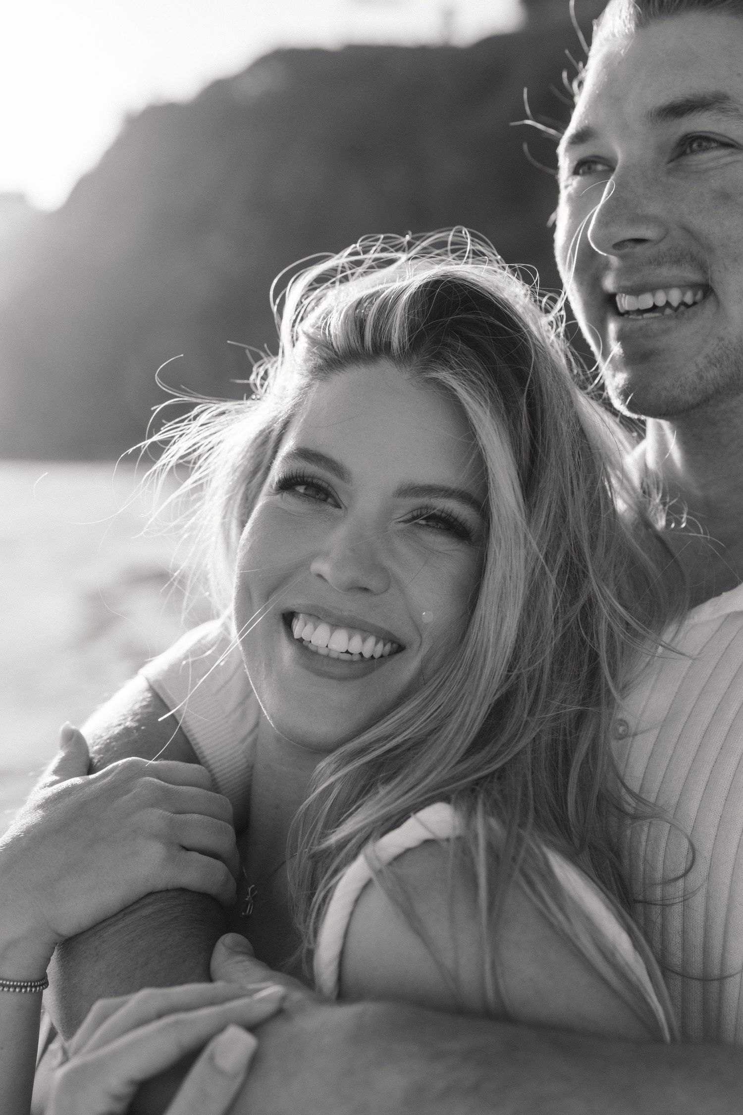 Black and white photo of happy couple showing off engagement ring while embracing on beach.