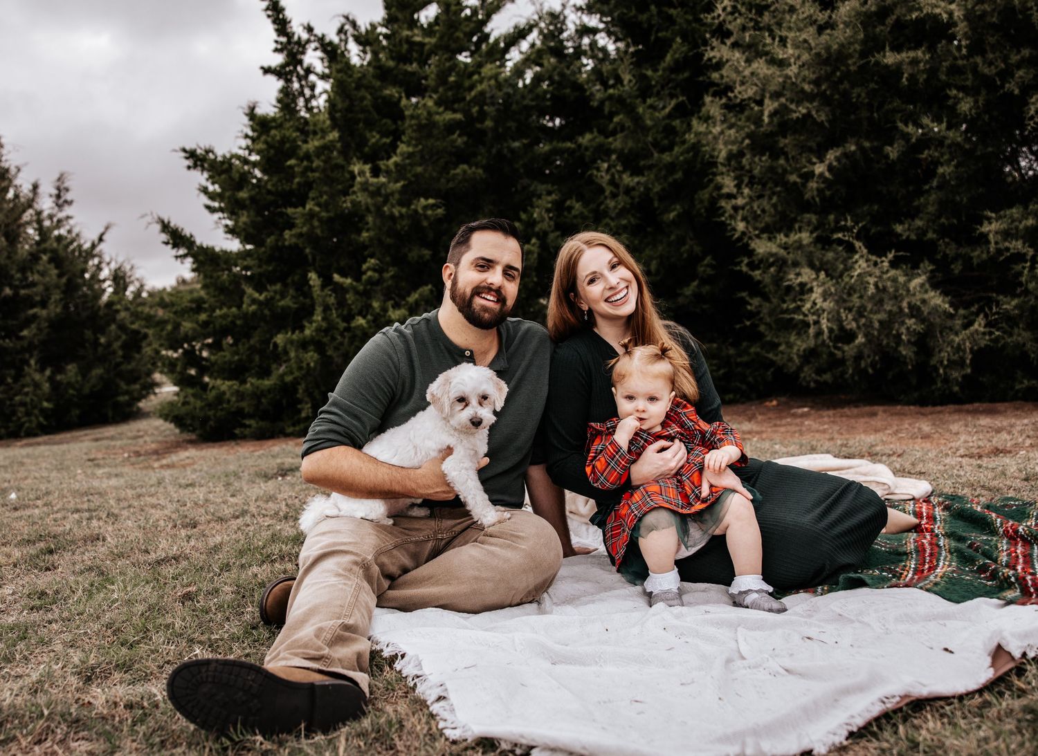 Happy family with a white dog and baby sitting on blanket outdoors near evergreen trees.