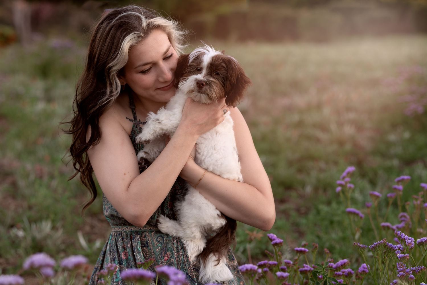 Person in a floral field holding and cuddling with a small fluffy dog among purple wildflowers.