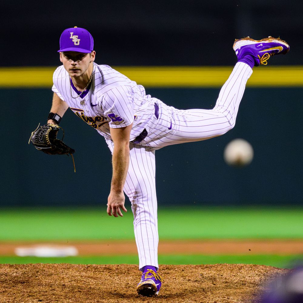 Baseball pitcher in purple and white pinstriped uniform follows through on delivery during night game.