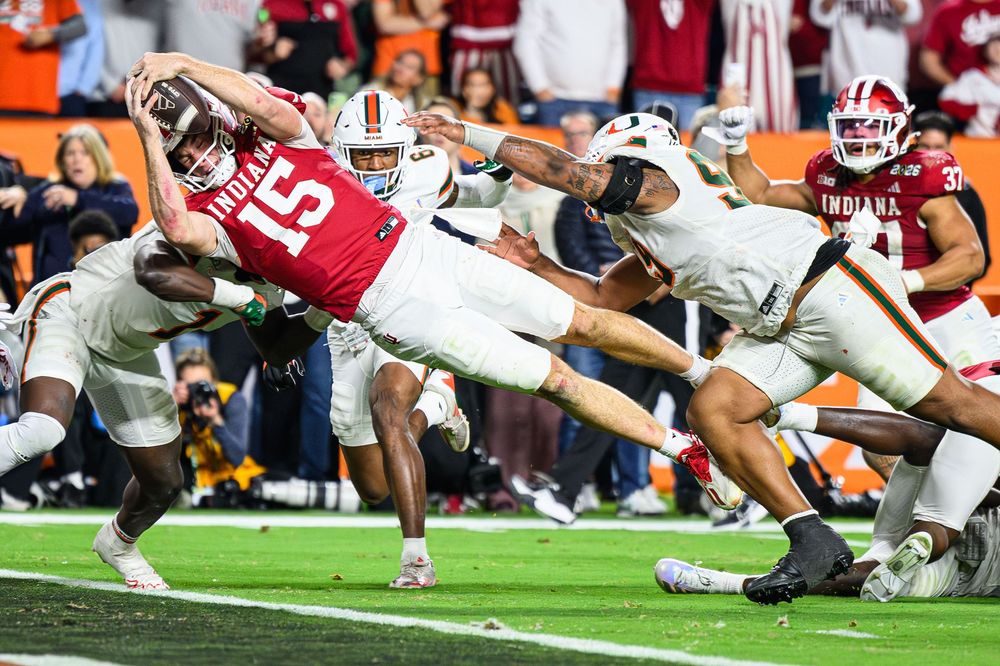 College football player in red and white uniform dives horizontally to score a touchdown during intense game action.