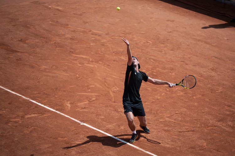 Tennis player serving on clay court captured against harsh shadows in dramatic lighting.