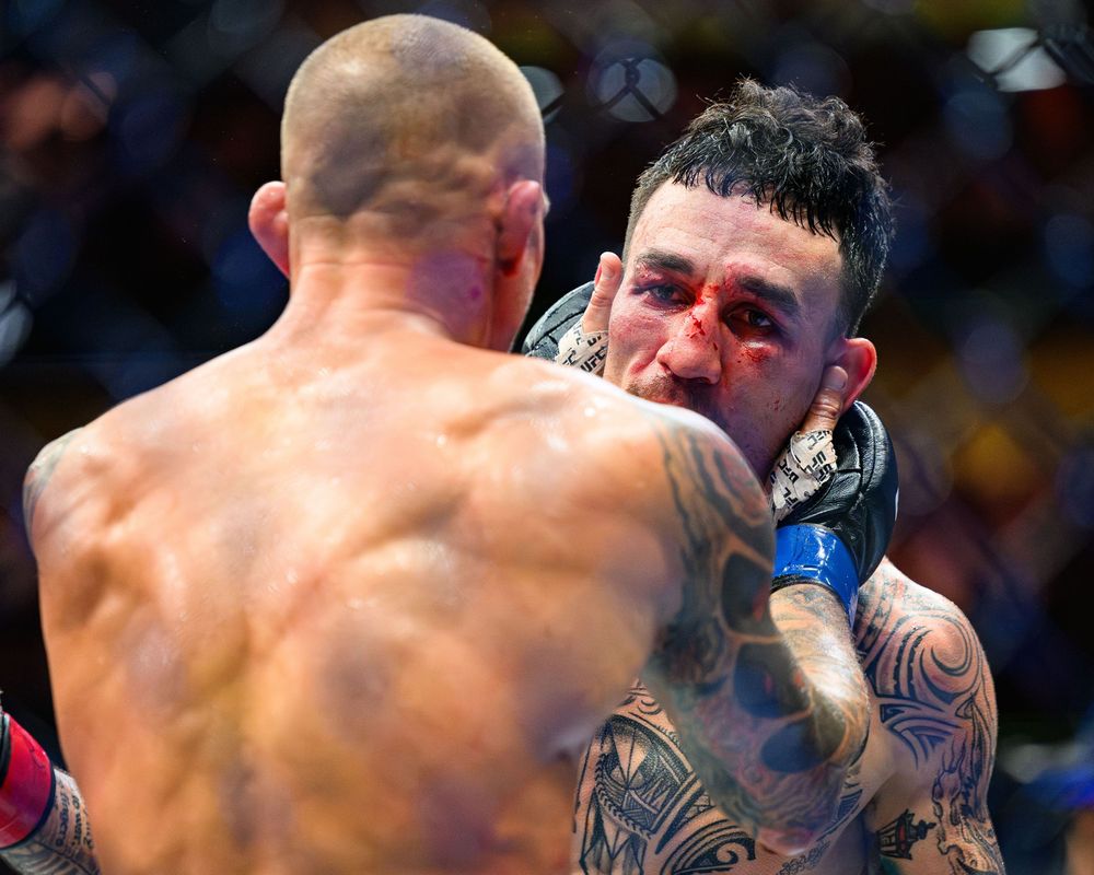 A tattooed MMA fighter bleeds from the face during an intense mixed martial arts bout in an octagon arena.