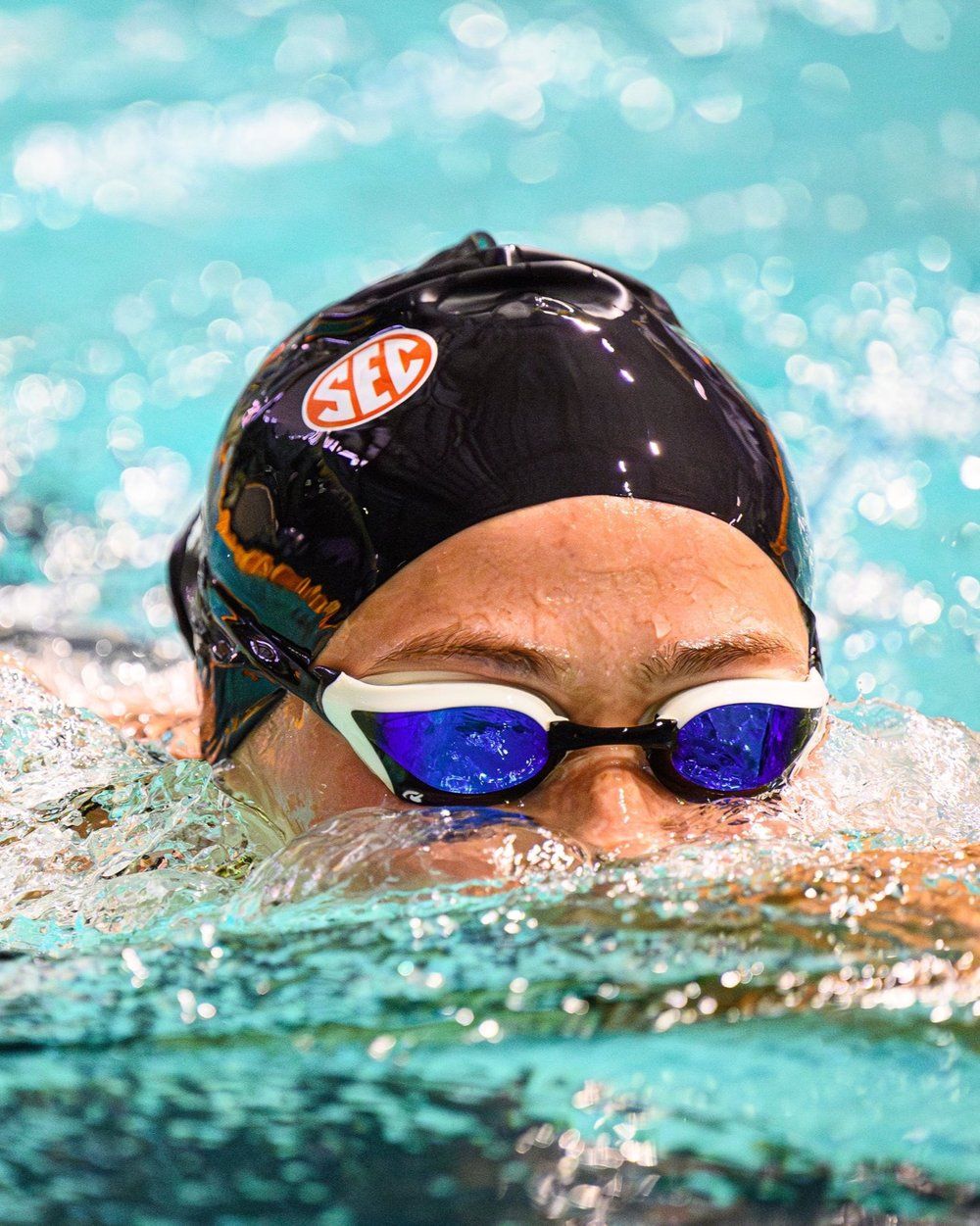 Swimmer in black cap and blue reflective goggles emerges from turquoise pool water.