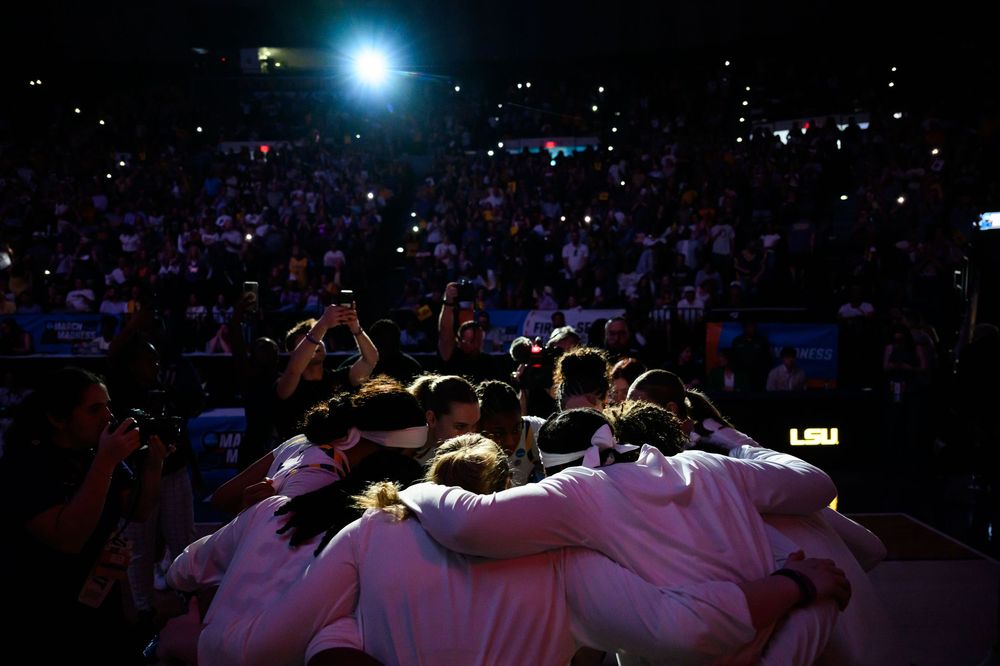 Sports team huddles together in a dark arena with crowd and bright lights in the background.