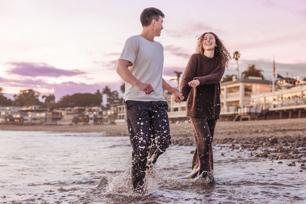 Couple splashing and laughing together in ocean waves at sunset on a beach with coastal homes in background.