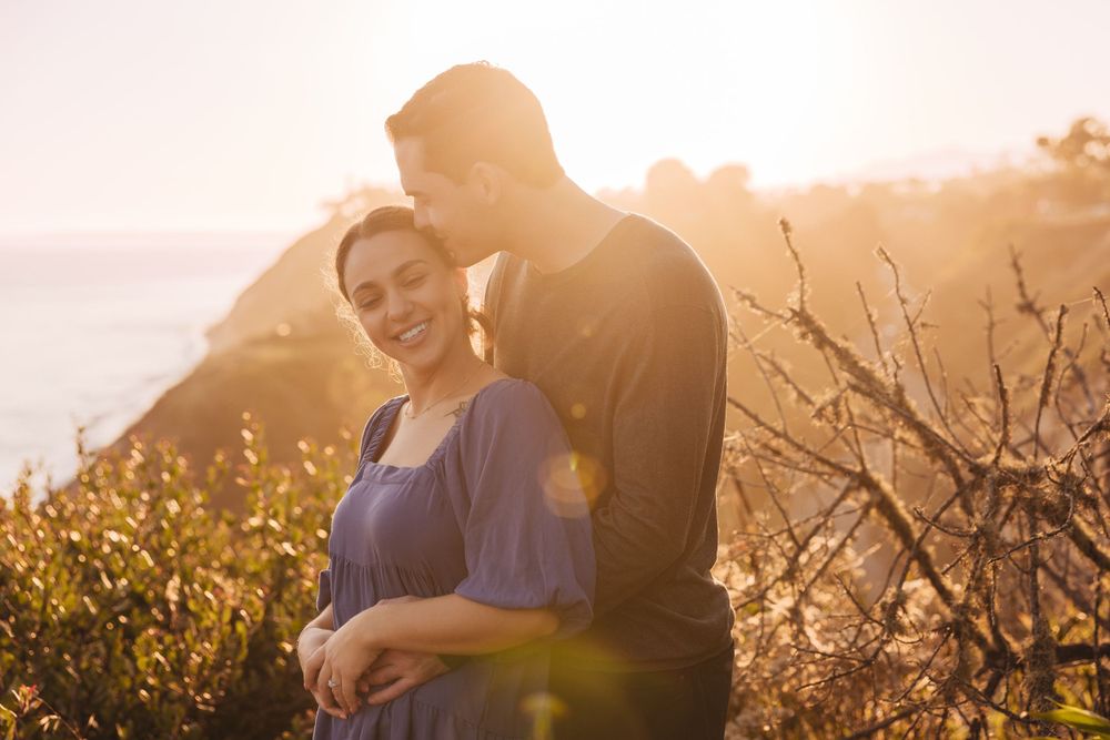 A romantic couple embraces at golden hour against a scenic coastal landscape with warm sunlit bushes.