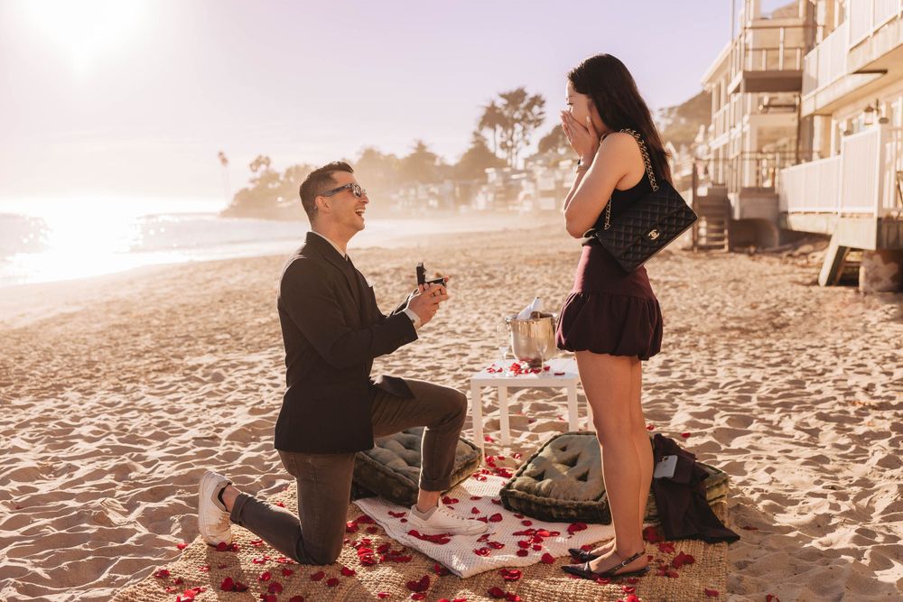 Romantic beach proposal scene at sunset with rose petals scattered on the sand near oceanfront homes.