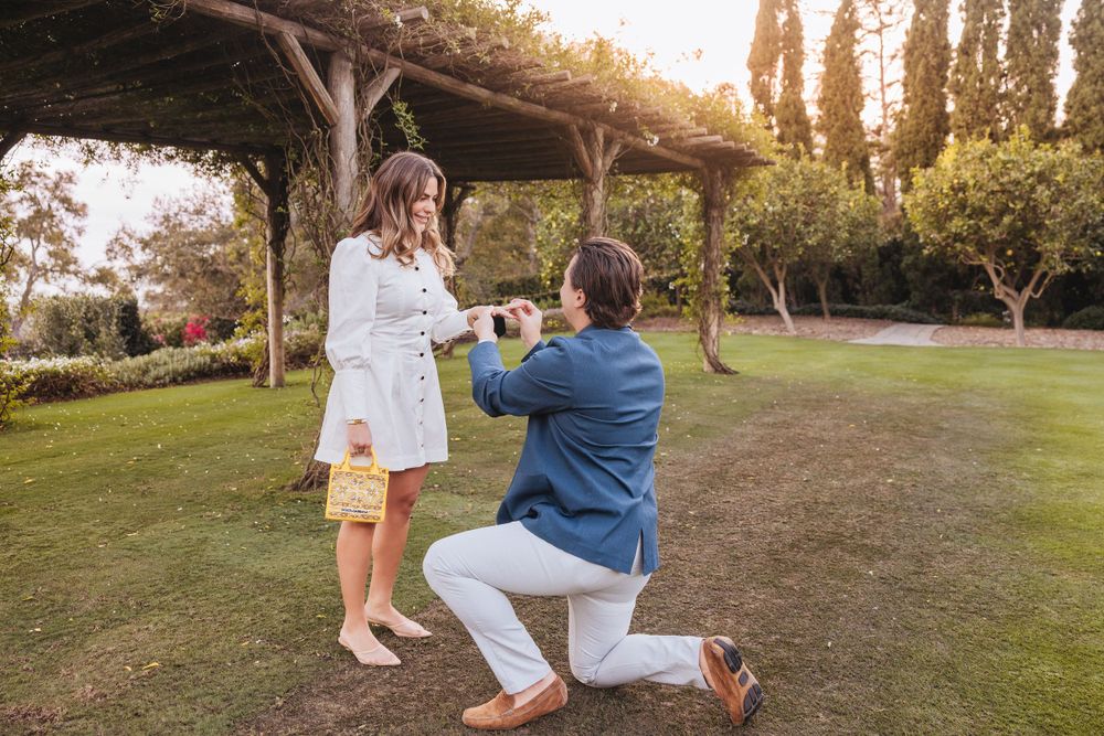 A romantic proposal moment unfolds under a rustic wooden pergola at sunset in a garden setting.