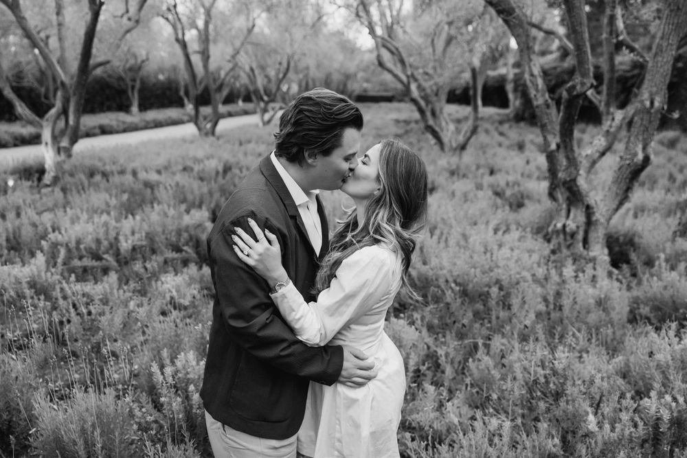 A romantic black and white photograph of a couple embracing in a garden setting surrounded by trees and flowers.