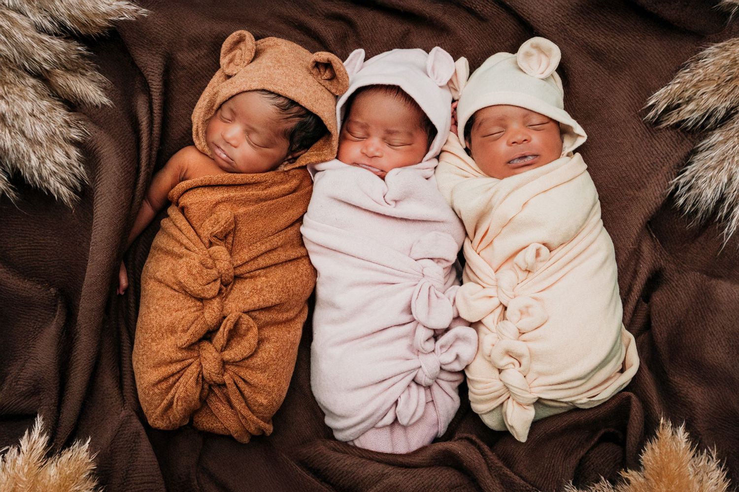 Three sleeping newborn babies swaddled in brown, pink, and cream colored wraps lying together on a brown blanket. 3 little bears. Triplets